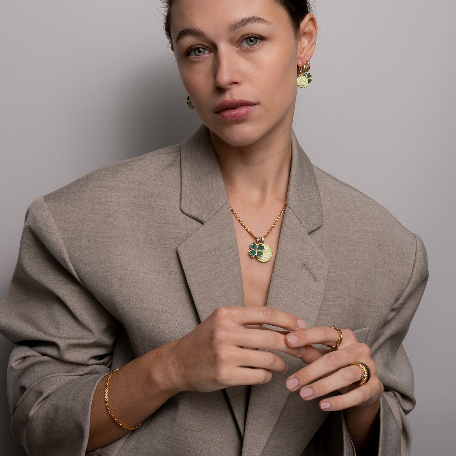 Frau in taupefarbenem Blazer trägt goldenen Schmuck mit grünen Akzenten, darunter die Ti Sento Milano Halskette - The Good Luck und passende Halskette, Ohrringe, Ringe und Armband von Ti Sento Milano.