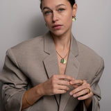Frau in taupefarbenem Blazer trägt goldenen Schmuck mit grünen Akzenten, darunter die Ti Sento Milano Halskette - The Good Luck und passende Halskette, Ohrringe, Ringe und Armband von Ti Sento Milano.