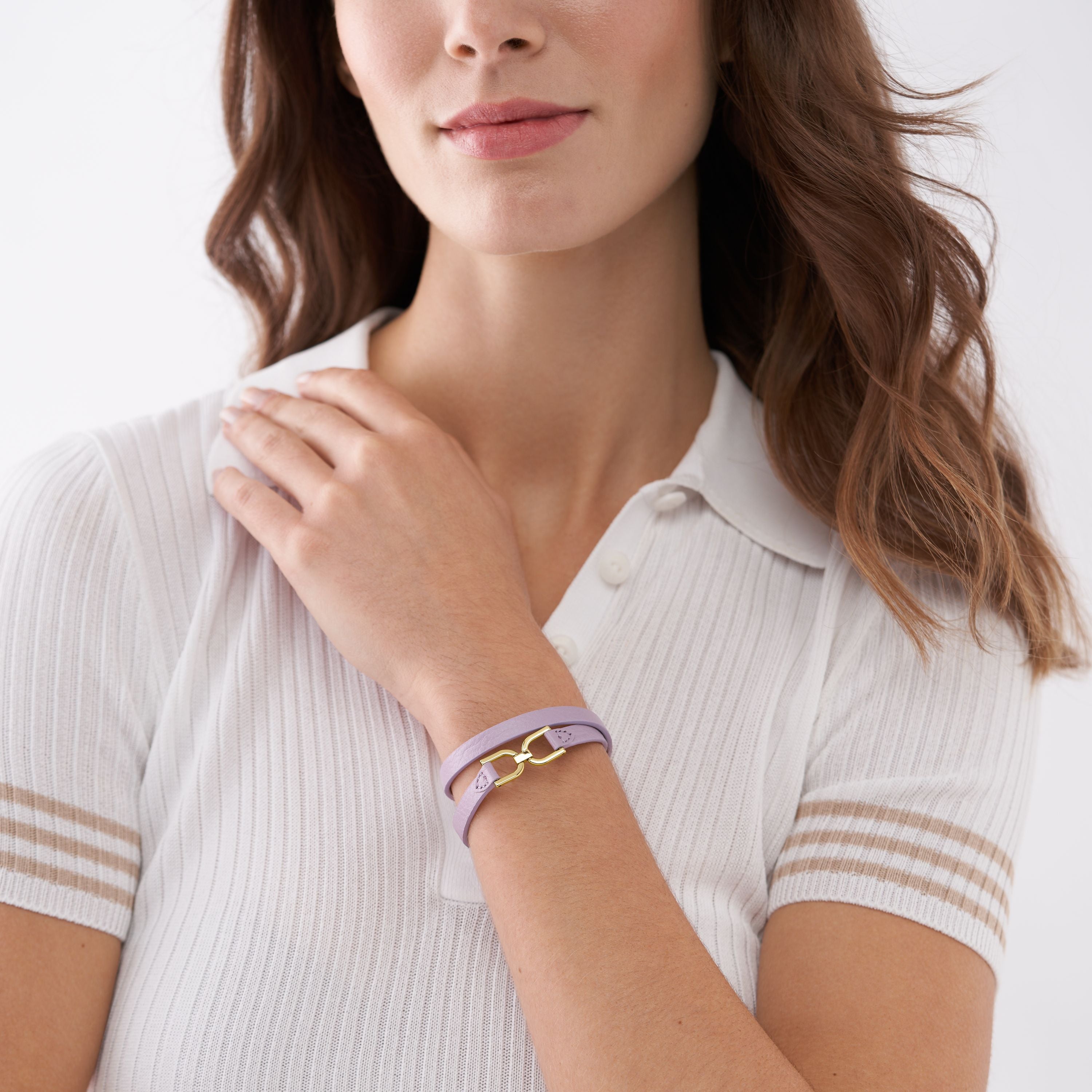 Eine Frau mit weißem Kragenhemd berührt ihre Schulter und präsentiert das Fossil Armband - Heritage D-Link Lavendel von Fossil - ein elegantes Armband mit goldenen Akzenten, das eine subtile Ergänzung für jede Schmuckkollektion darstellt.