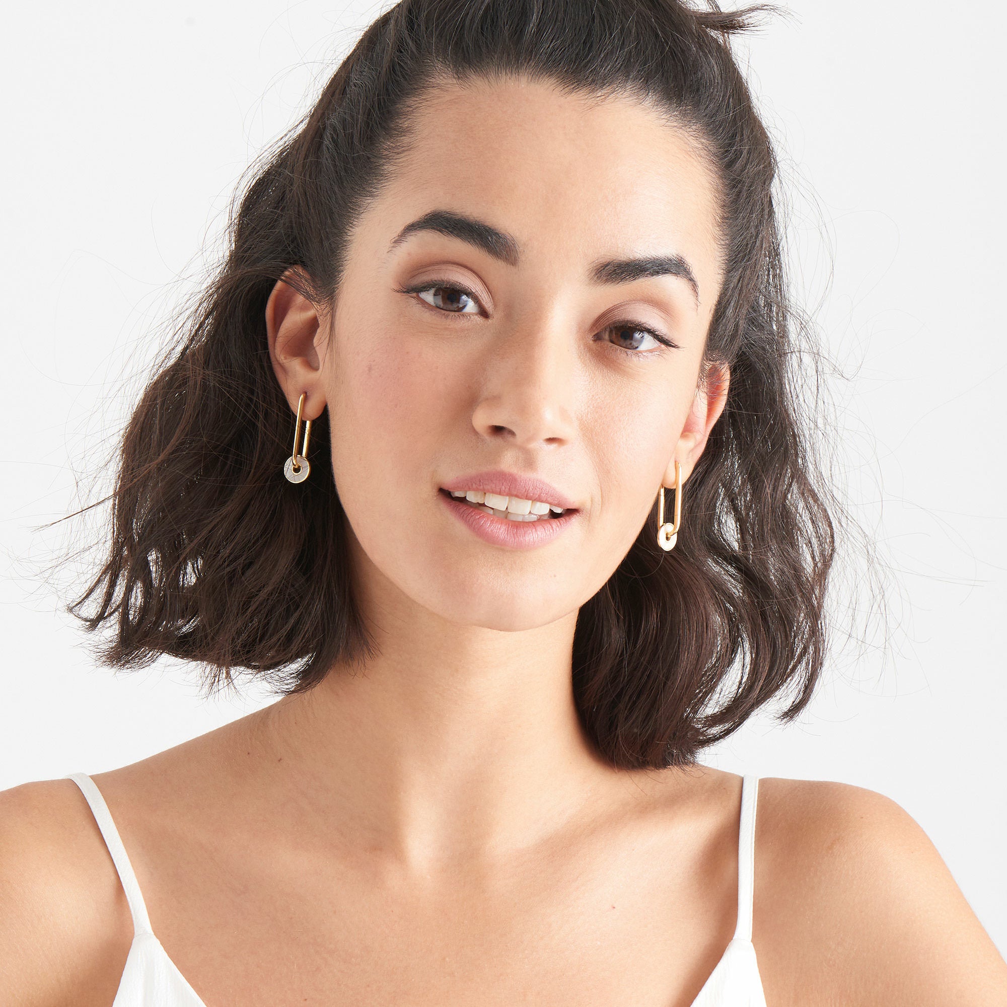 Frau mit schulterlangem dunklem Haar, goldenen Ohrringen und weißem Mieder lächelt in die Kamera. Sie vervollständigt ihren Look mit Ania Haie Ohrhänger - White Hidden Gem von Ania Haie, einem eleganten Blickfang, der die Aufmerksamkeit auf sich zieht.