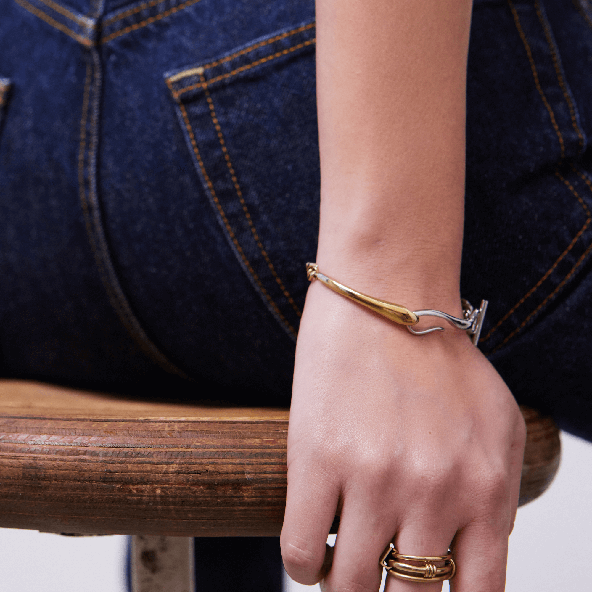 Goldenes Armband und Ringe an einer Hand, lässig auf einem Holzstuhl vor Jeans.