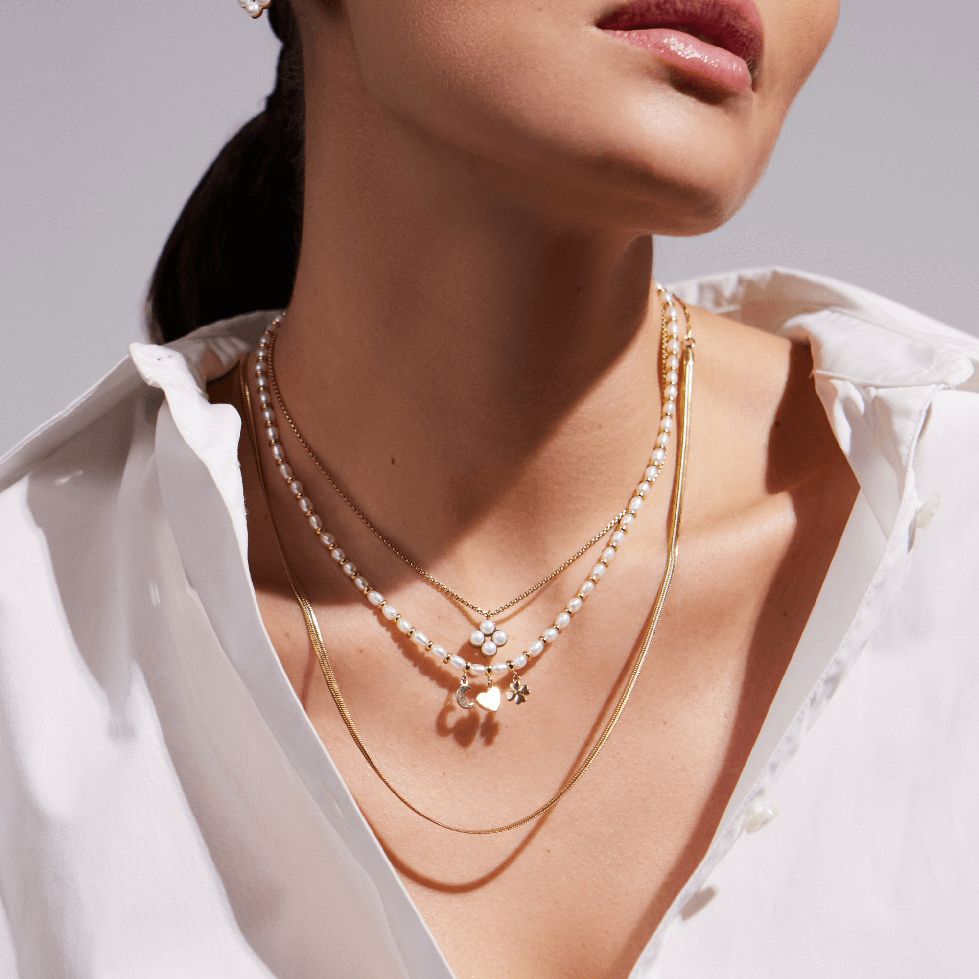 Frau mit offenem weißen Hemd trägt mehrere goldene Halsketten mit Perlen und Anhängern.