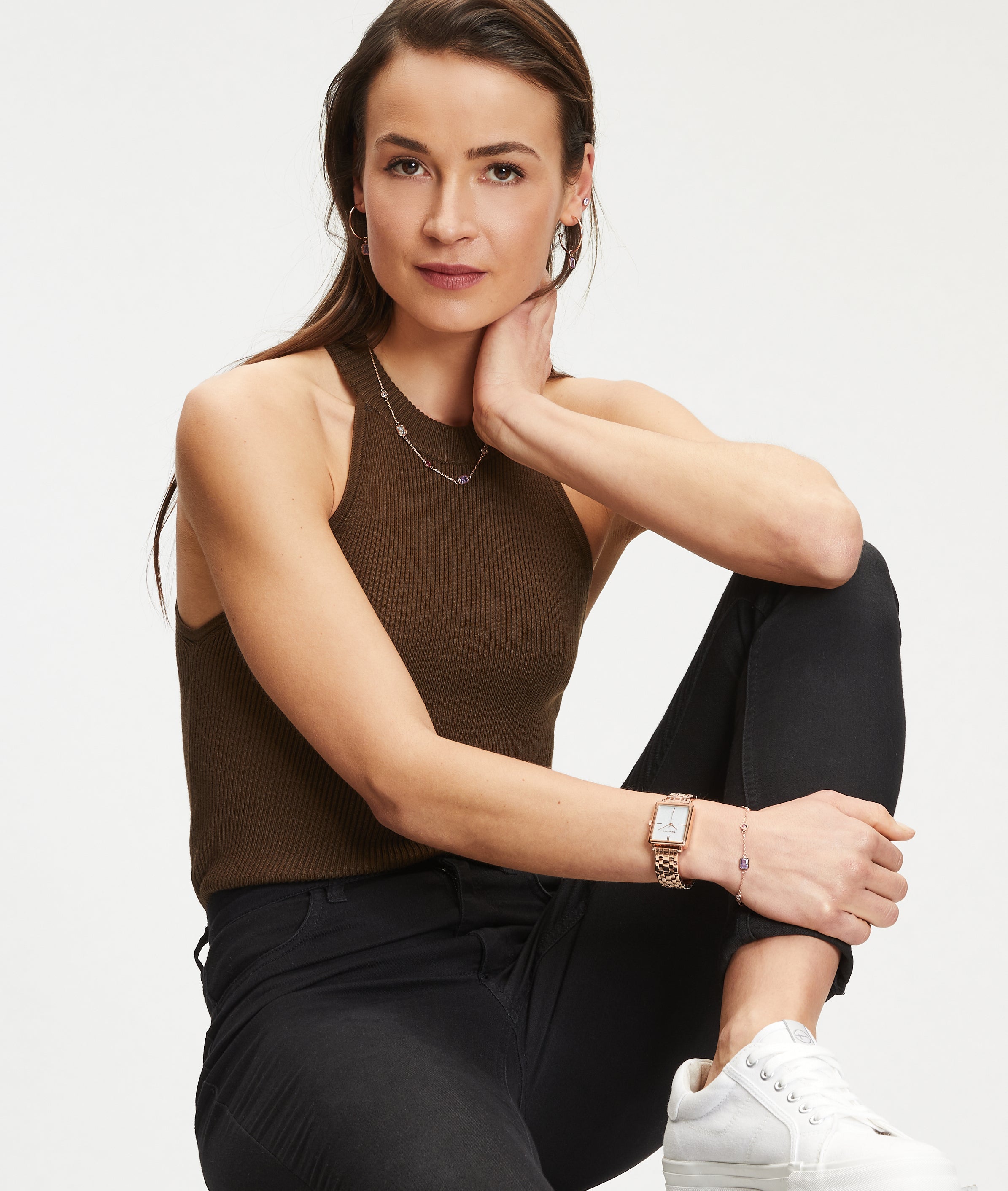 Eine Frau in einem braunen ärmellosen Oberteil und schwarzen Jeans sitzt mit einem Knie hoch, trägt weiße Turnschuhe und Tamaris Ohrstecker - Baguette by Tamaris, die ihrem Look einen eleganten Touch von Ohrschmuck verleihen.