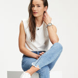 Eine Frau in einem lässigen weißen ärmellosen Oberteil und blauen Jeans sitzt im Schneidersitz und schaut in die Kamera. Sie trägt die Tamaris Armbanduhr - The Basic Must-Have, die ihrem lässigen Stil Raffinesse verleiht.