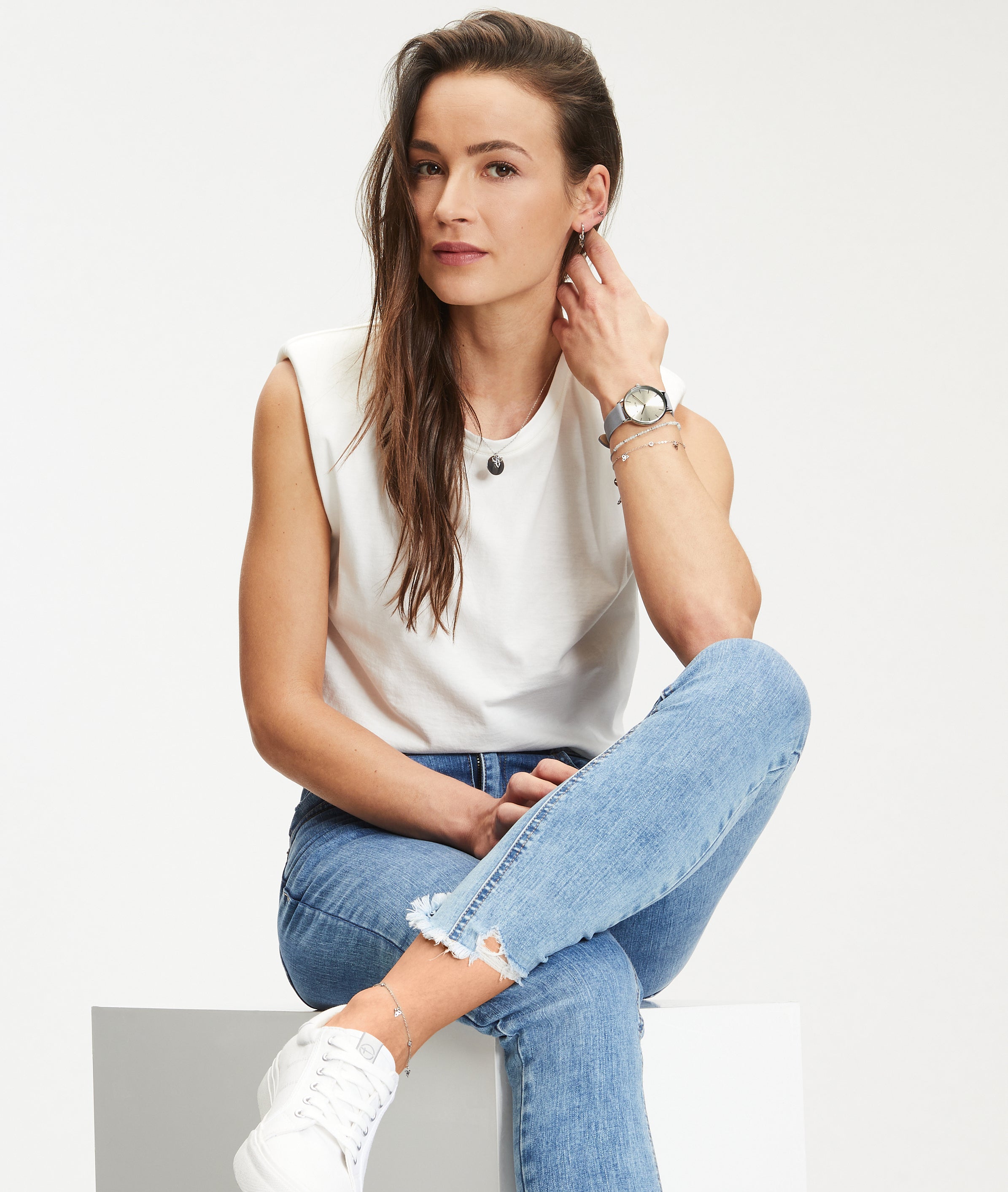 Eine Frau in einem lässigen weißen ärmellosen Oberteil und blauen Jeans sitzt im Schneidersitz und schaut in die Kamera. Sie trägt die Tamaris Armbanduhr - The Basic Must-Have, die ihrem lässigen Stil Raffinesse verleiht.