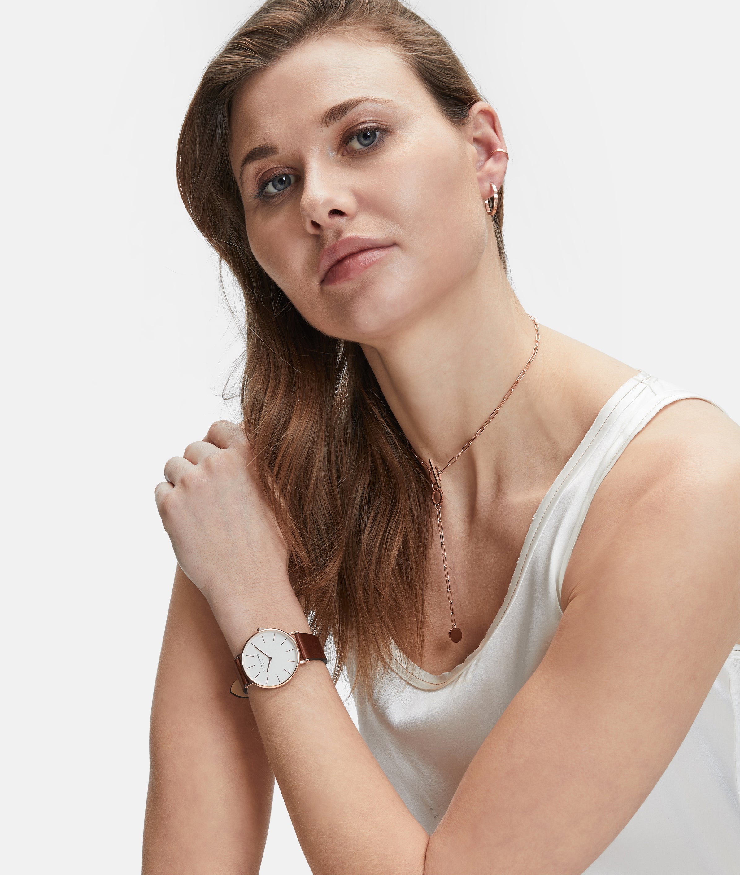 Frau in einem weißen Tank-Top, die eine Armbanduhr, mehrere Ketten und die LIEBESKIND BERLIN Ear Cuff - The Small Basic Logo by Liebeskind Berlin trägt und in die Kamera schaut.