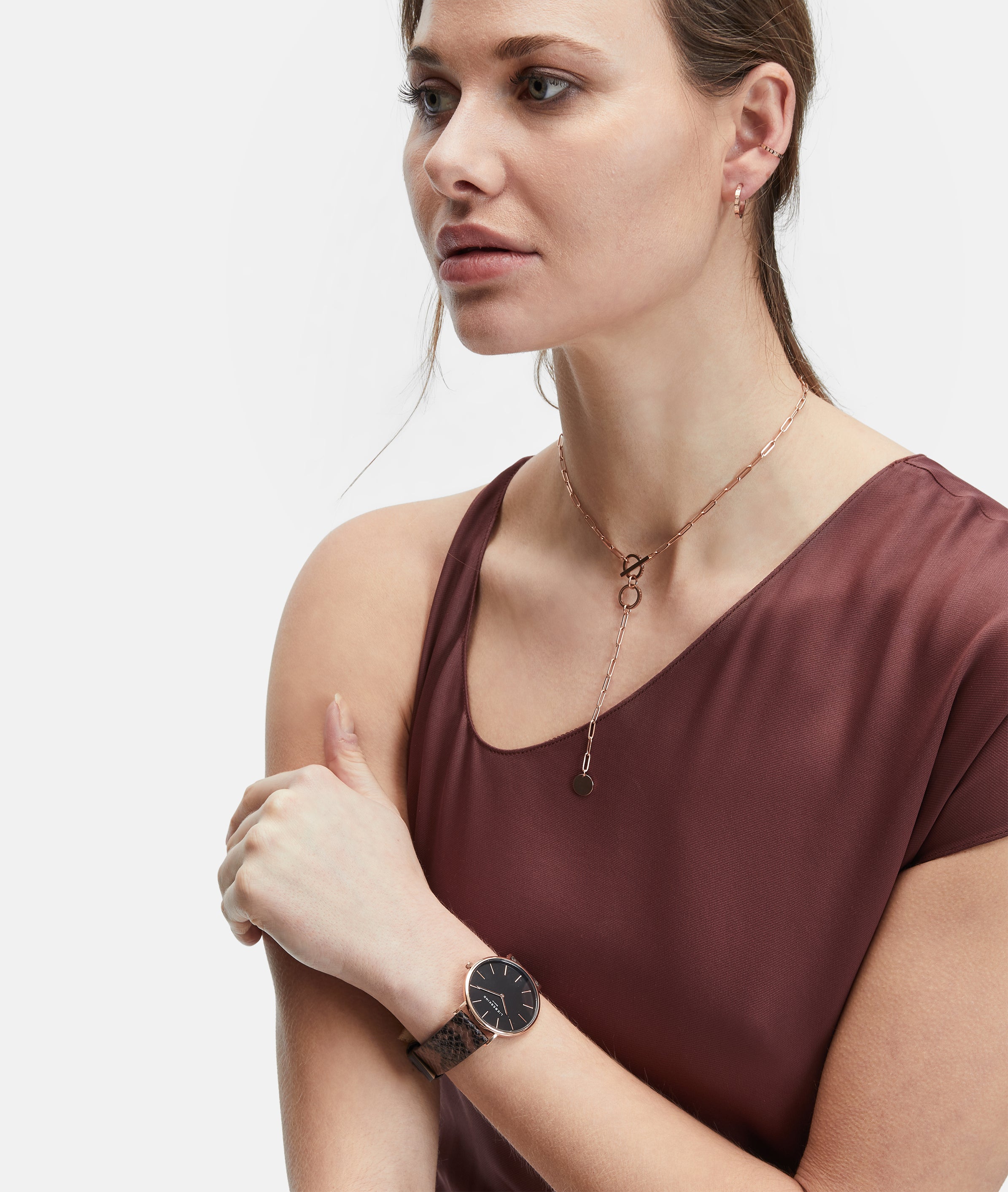 Frau in einem ärmellosen weinroten Oberteil, trägt eine Halskette, eine elegante schwarze Armbanduhr und die LIEBESKIND BERLIN Ear Cuff - The Small Basic Logo by Liebeskind Berlin, mit Blick zur Seite.
