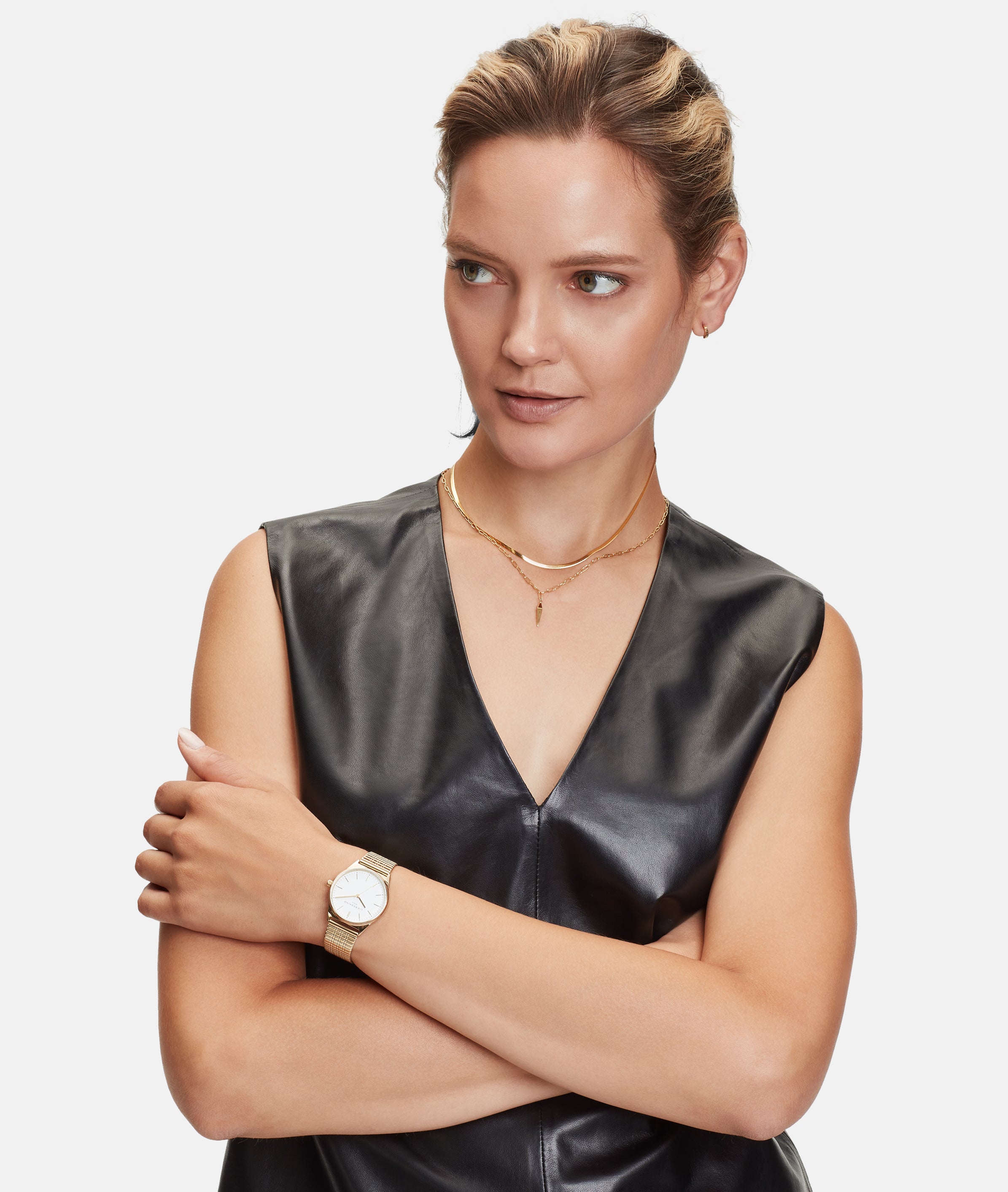 Eine Frau in einem schwarzen ärmellosen Ledertop verschränkt die Arme und trägt mehrere goldene Halsketten, darunter die Liebeskind Berlin Halskette - The Sleek, sowie eine goldene Uhr, um einen schicken und modernen Look zu kreieren.