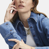 Frau in Jeanshemd und Jeans, trägt Tamaris Creole - Coin Ohrringe von Tamaris, posiert mit der Hand vor dem Gesicht vor weißem Hintergrund.