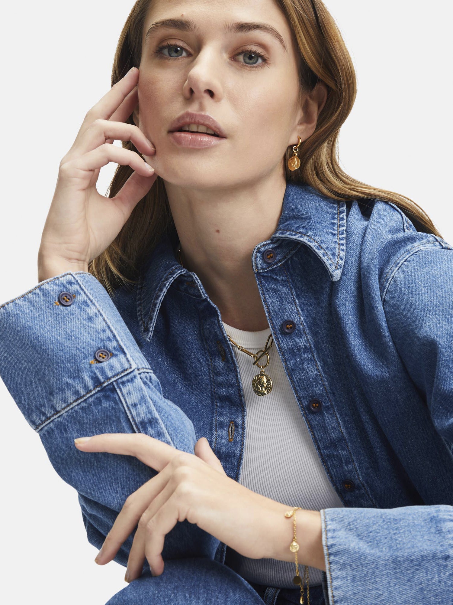 Frau in Jeanshemd und Jeans, trägt Tamaris Creole - Coin Ohrringe von Tamaris, posiert mit der Hand vor dem Gesicht vor weißem Hintergrund.
