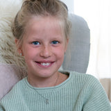Junges Mädchen mit blonden Haaren und blauen Augen lächelt im Haus. Sie trägt einen hellgrünen Pullover und die Cool Time Kids Halskette - The Cool Bully von Cool Time.