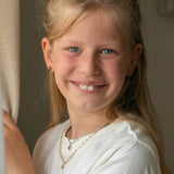 Lächelndes junges Mädchen mit blonden Haaren und blauen Augen trägt ein weißes Hemd und mehrlagige goldene Halsketten, mit der Cool Time Kids Halskette - The Cool Heart von Cool Time.