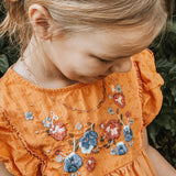 Ein junges Mädchen in einem orangefarbenen Kleid mit blauer und roter Blumenstickerei lächelt sanft und trägt die Cool Time Kids Halskette - The Cool Mini Link von Cool Time.
