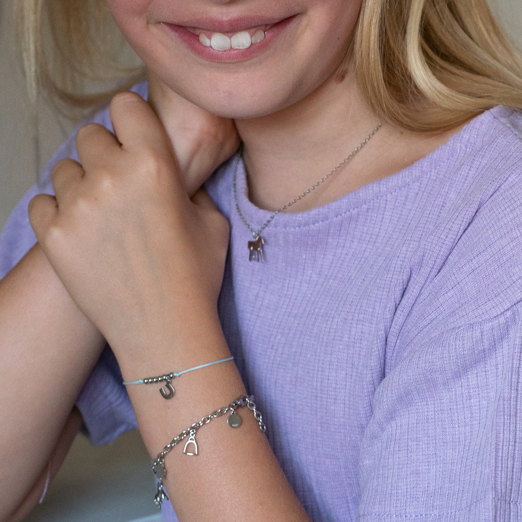 Lächelndes Kind in einem lavendelfarbenen Oberteil, das eine Halskette und das Cool Time Kids Armband - The Cool Charm von Cool Time trägt, das mit silbernen Anhängern verziert ist.
