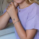Frau im lavendelfarbenen Hemd trägt das Cool Time Kids Armband - The Cool Charm von Cool Time und eine Halskette und stützt ihre Arme auf eine weiße Fläche - eine elegante Mischung aus Schmuck und Modeaccessoire.