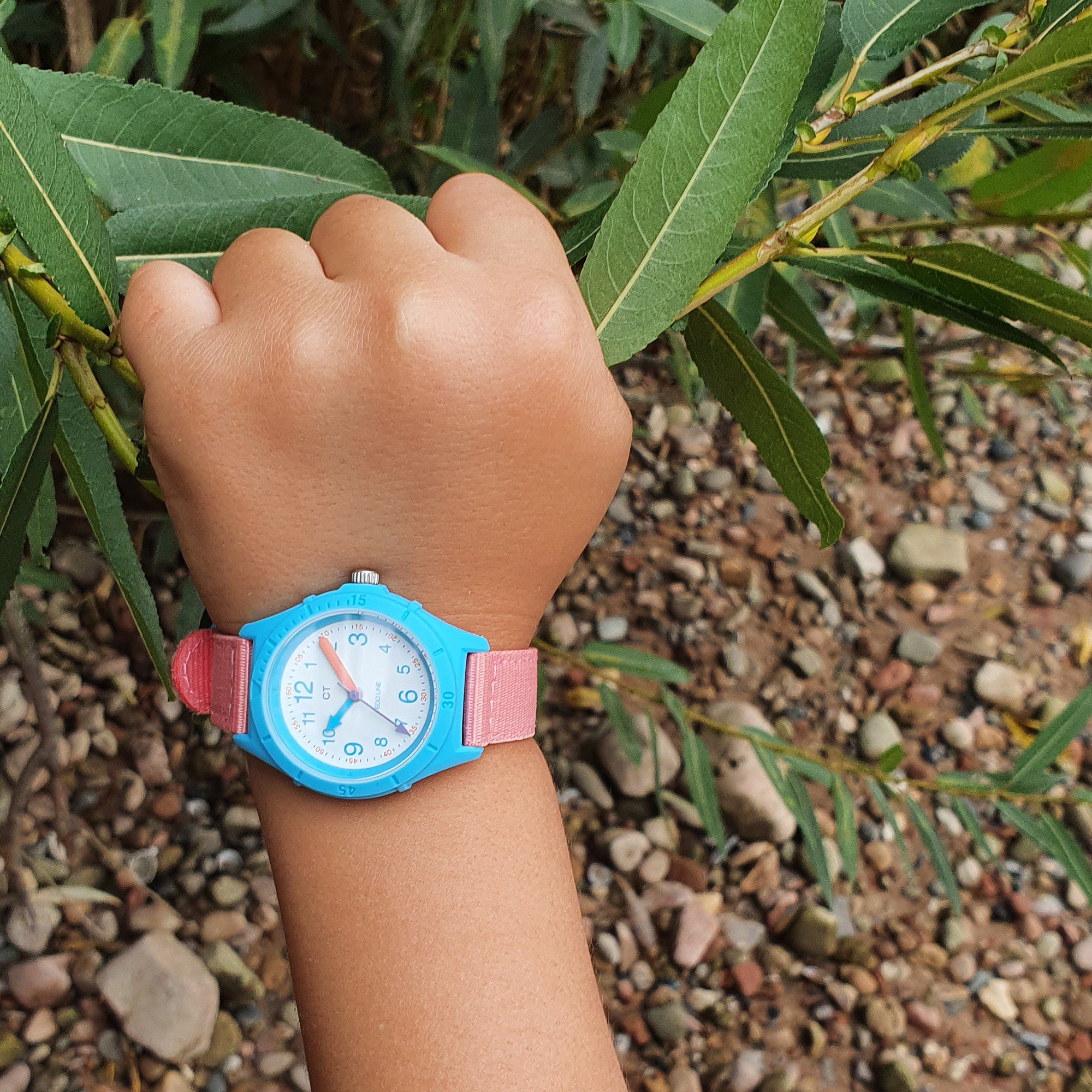 Das Handgelenk eines Kindes, das die Cool Time Kids Armbanduhr - The Cool Sustainable von Cool Time trägt, die aus recyceltem Ozeanplastik hergestellt ist, hält einen grünen Zweig über felsigem Grund.