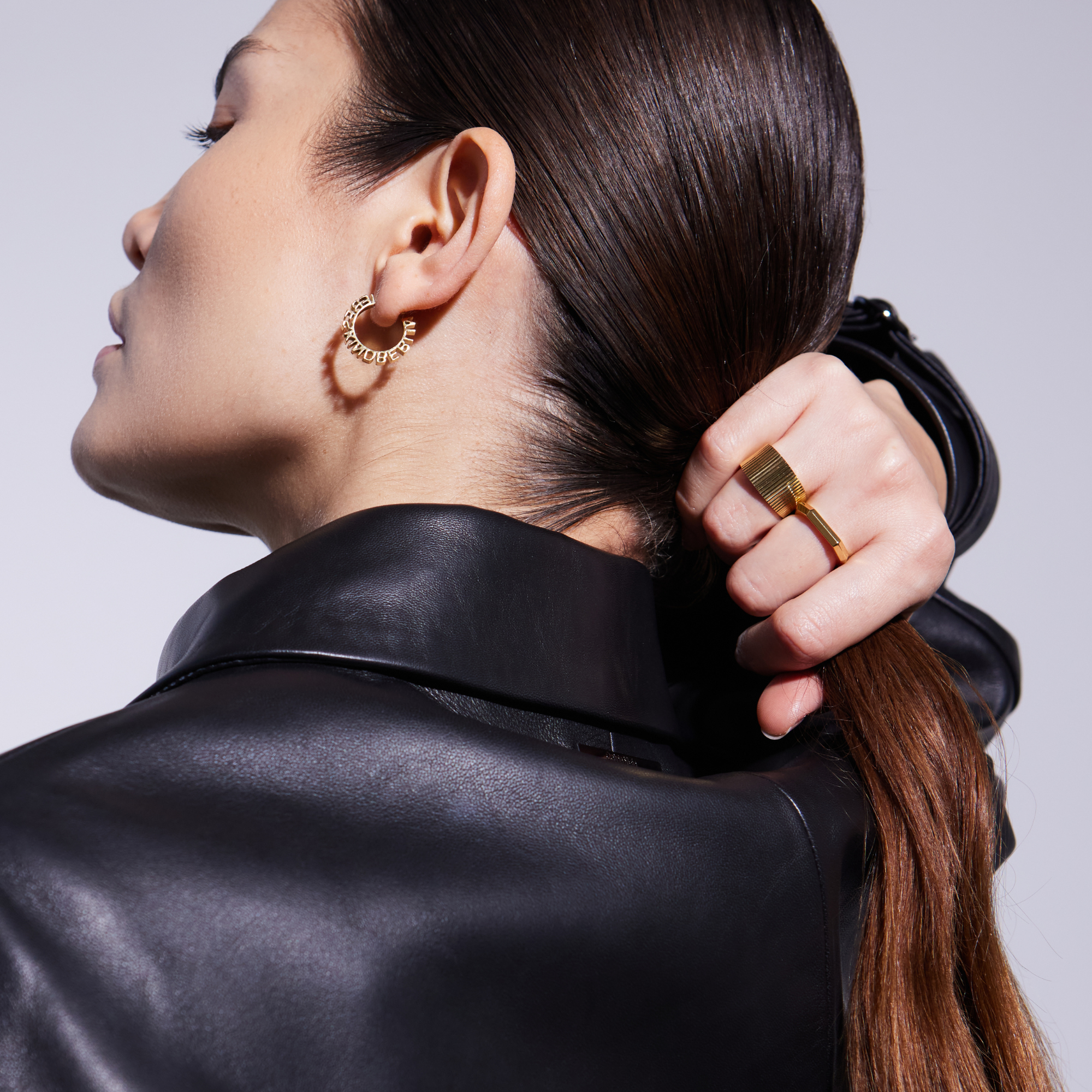 Frau in schwarzer Lederjacke hält Pferdeschwanz und trägt goldene Creolen und auffälligen Ring.