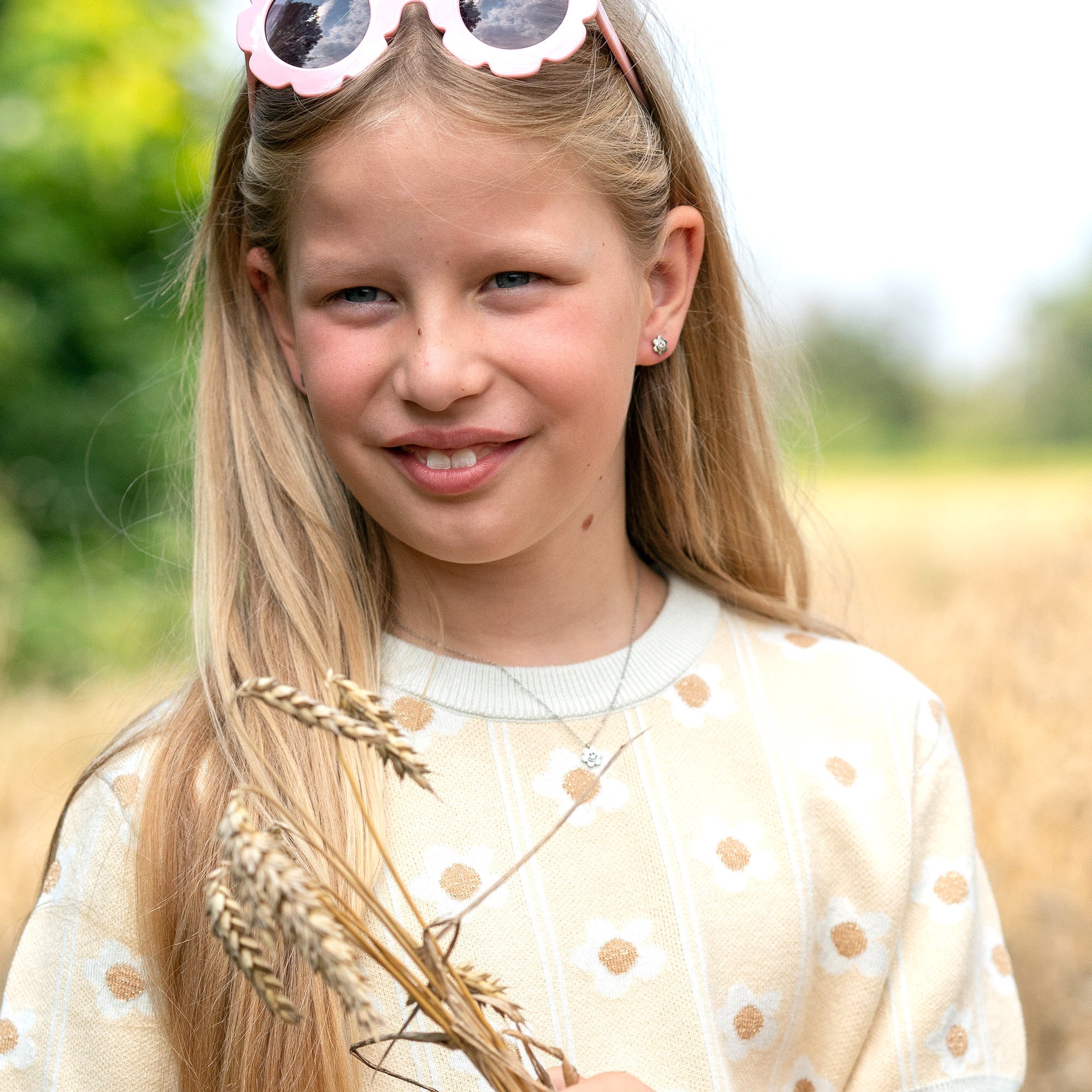 Lächelndes Mädchen mit langen blonden Haaren, das Weizenstängel hält, einen cremefarbenen Pullover und eine rosafarbene Sonnenbrille trägt, ausgestattet mit der Cool Time Kids Halskette - The Cool Smiley von Cool Time als charmanter Schmuck.