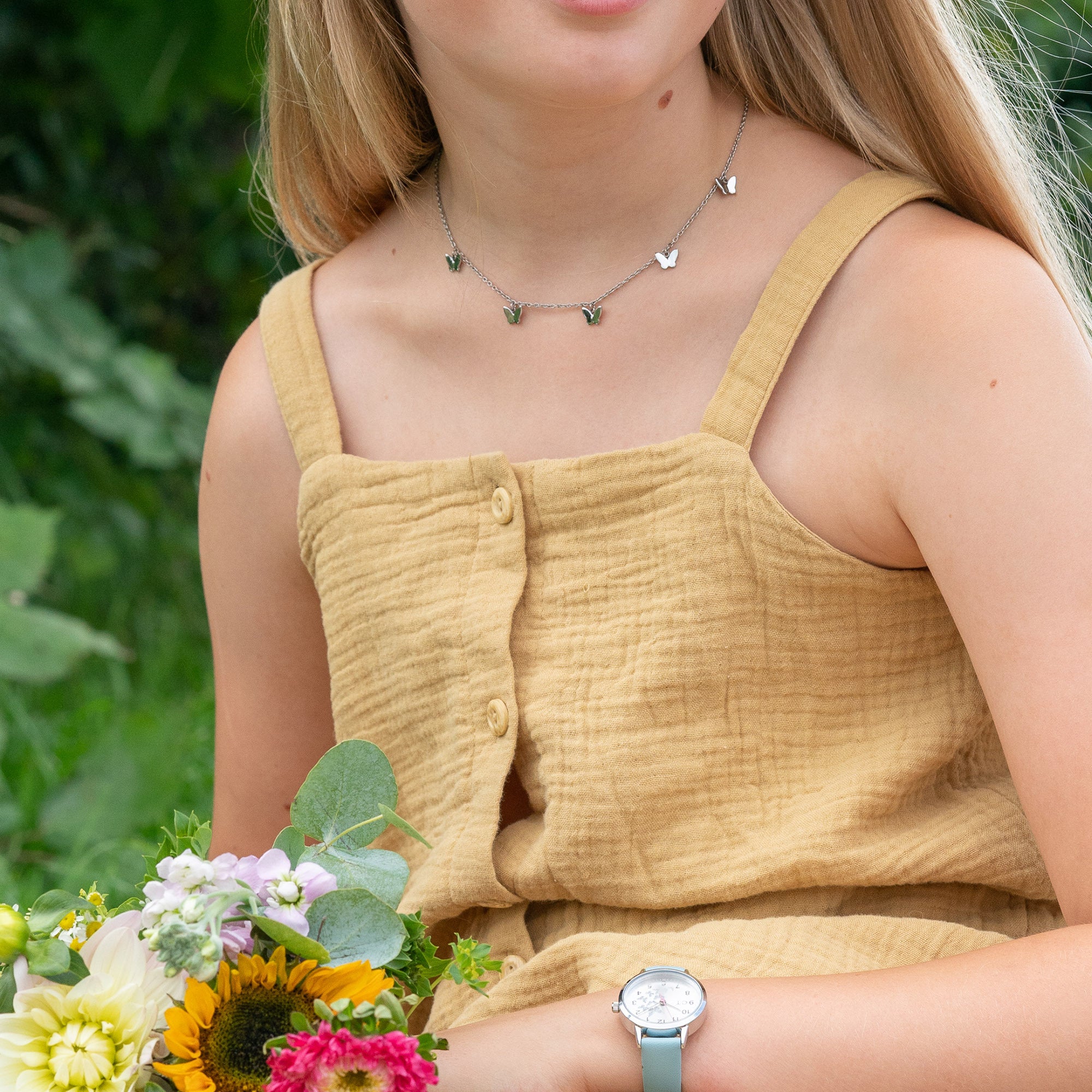 Mädchen im gelben Kleid, das einen Blumenstrauß in der Hand hält, trägt eine silberne Armbanduhr und die Cool Time Kids Halskette - The Cool Butterfly von Cool Time als Halskette.