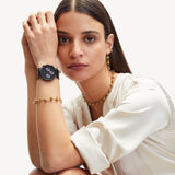 Frau in weißer Bluse trägt The Knot-Halskette von Liebeskind Berlin, Goldschmuck und eine schwarze Smartwatch und stützt ihren Arm auf ihr Knie.
