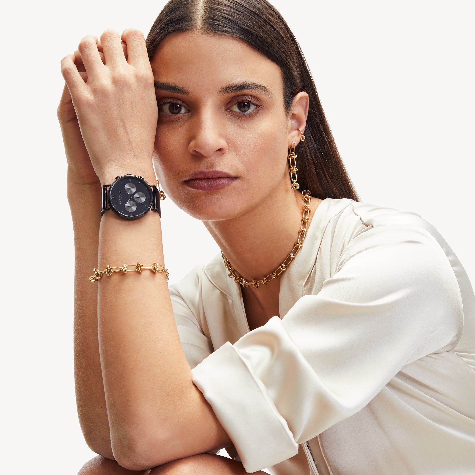 Frau in weißer Bluse trägt The Knot-Halskette von Liebeskind Berlin, Goldschmuck und eine schwarze Smartwatch und stützt ihren Arm auf ihr Knie.