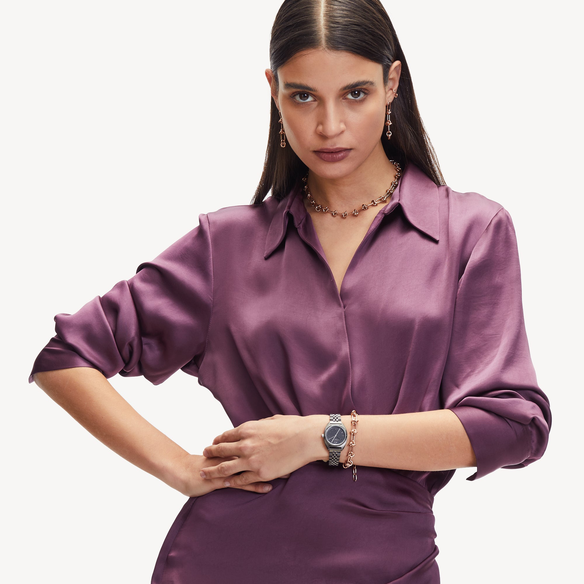 Eine Frau in einem lilafarbenen Seidenkleid posiert selbstbewusst mit der Liebeskind Berlin Halskette - The Knot und einer Uhr und setzt damit einen extravaganten Look vor einem schlichten Hintergrund in Szene.