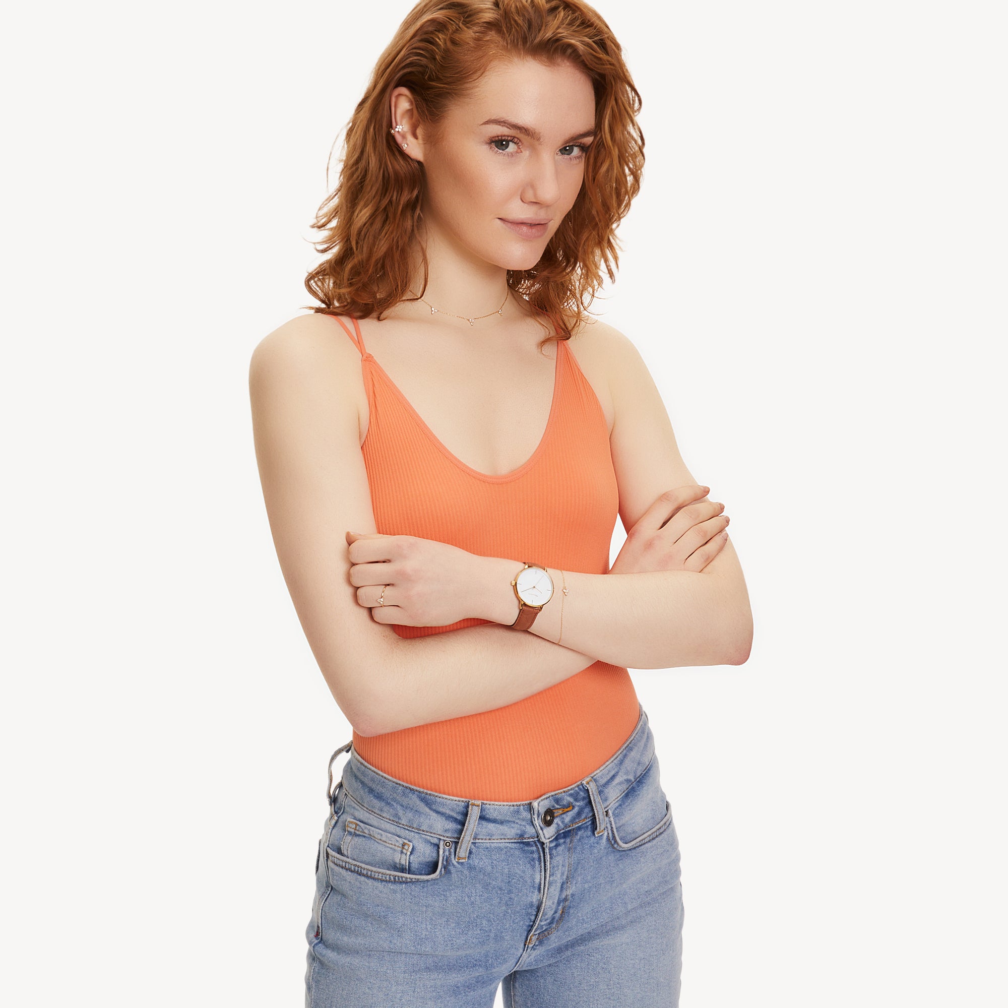 Frau mit gewellten roten Haaren in einem orangefarbenen Tank-Top und Jeans, Arme verschränkt, trägt ein Tamaris Armband - Sparkle Flowers und eine Uhr, auf weißem Hintergrund.