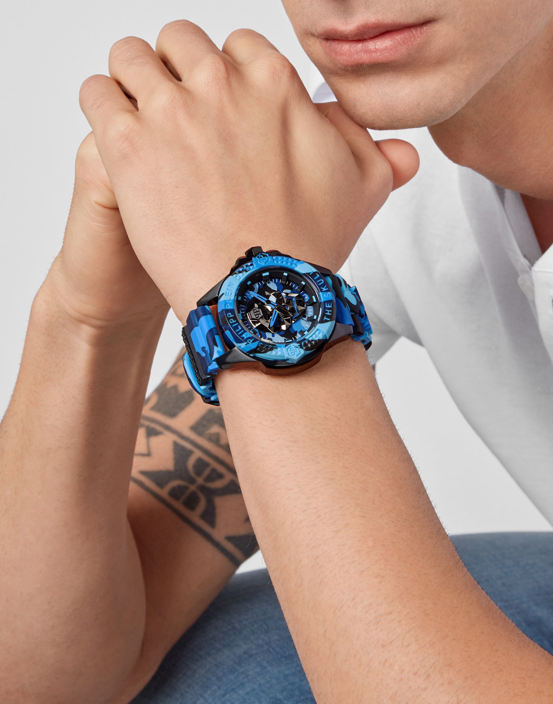 Eine Person mit gefalteten Händen trägt die Philipp Plein Armbanduhr - The Blue Camouflage und zeigt ein Unterarmtattoo.