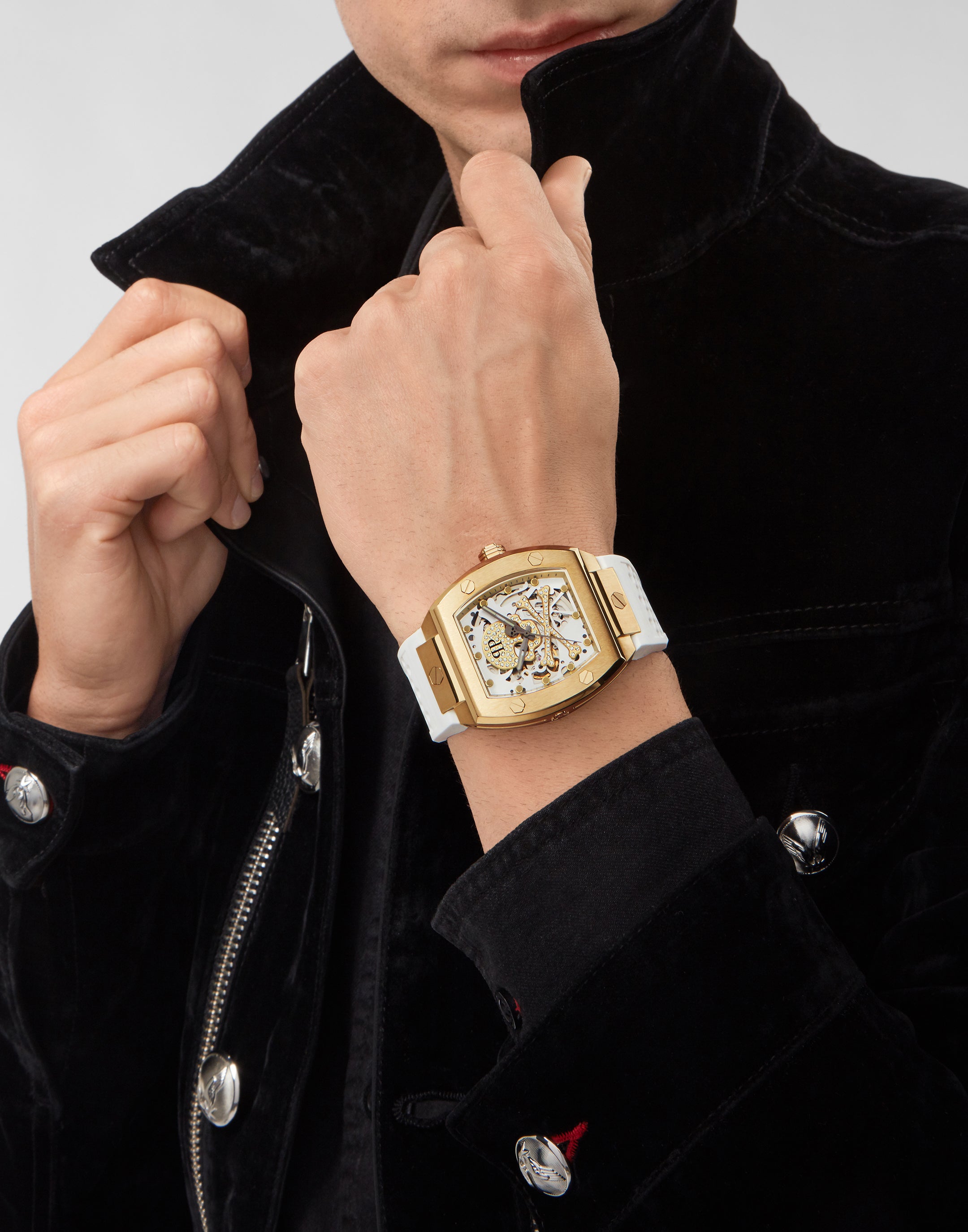 Eine Person in einem schwarzen Jackett rückt den Kragen zurecht und präsentiert eine Philipp Plein Armbanduhr - The White Golden $keleton mit einem großen goldenen Gehäuse und weißem Armband.
