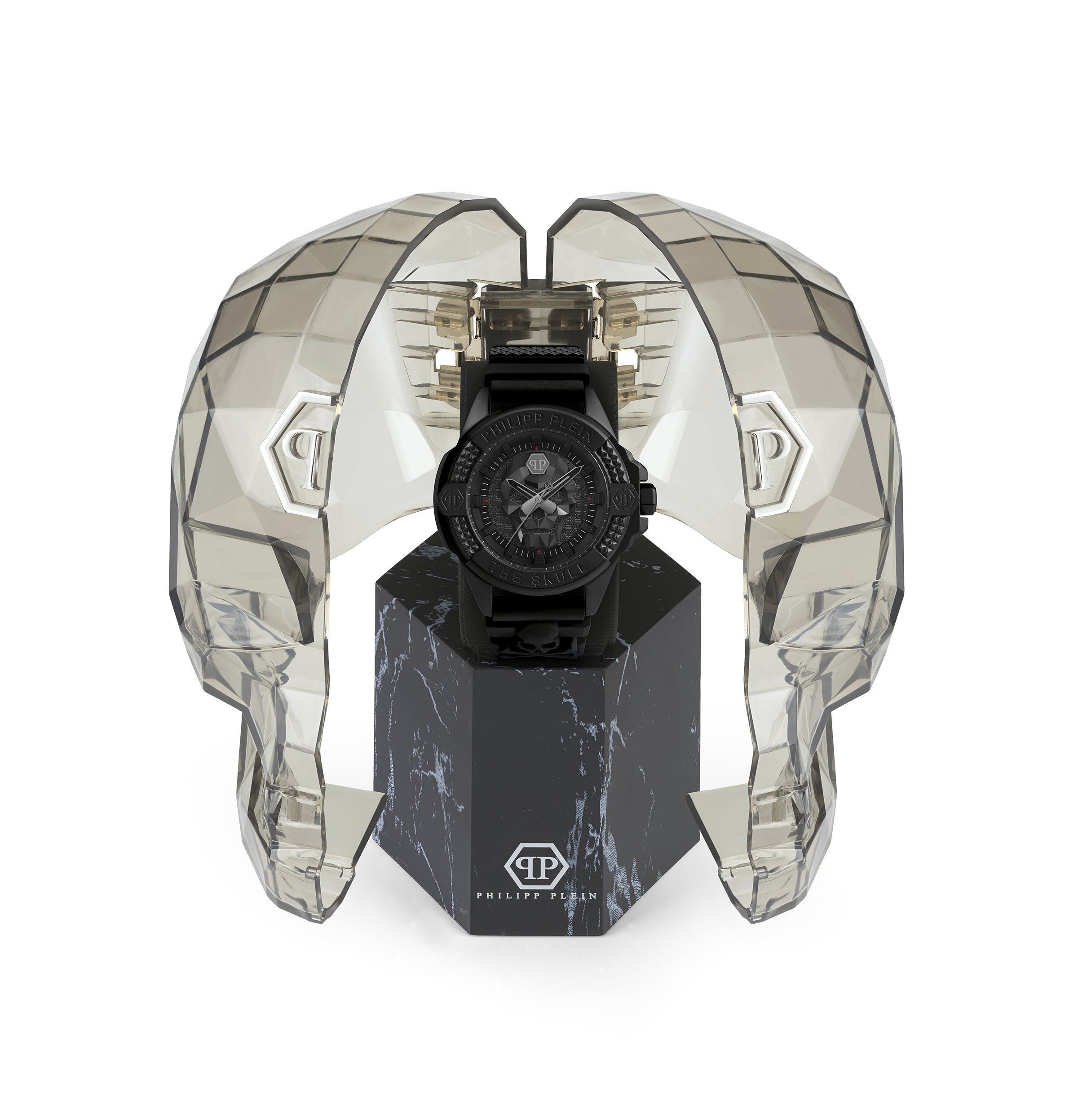 Die Philipp Plein Armbanduhr - The Solid Black $kull, eine Herrenuhr von Philipp Plein, wird in einem transparenten Gehäuse in Form eines Totenkopfes auf einem schwarzen Marmorständer präsentiert.