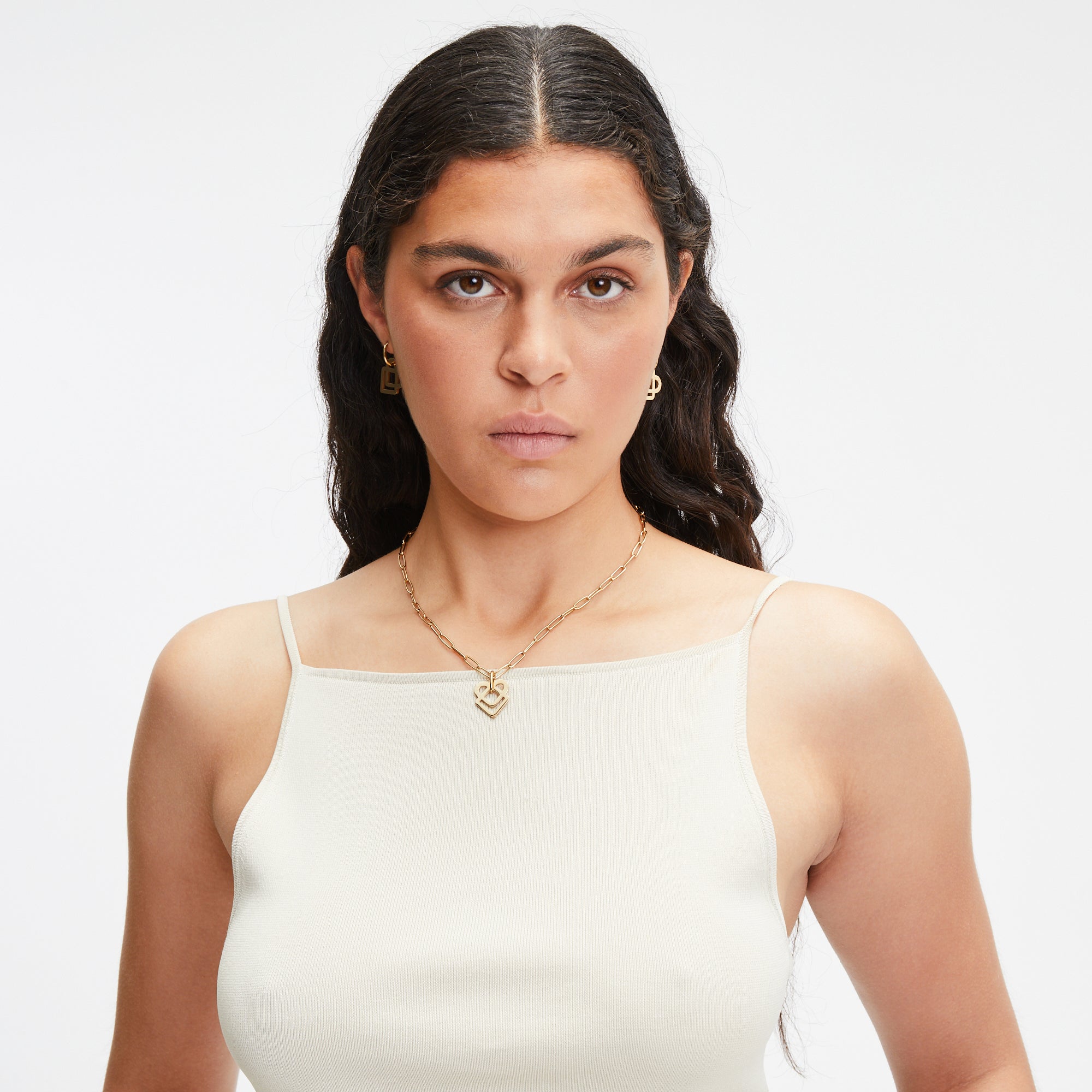 Eine Frau mit langen dunklen Haaren in einem weißen Tanktop trägt Goldschmuck und LIEBESKIND BERLIN Ohrhänger - Colorful Icon by Liebeskind Berlin und schaut ernst in die Kamera.