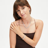 Eine Frau in einem braunen ärmellosen Kleid lächelt in die Kamera und trägt Tamaris Creole - Rocking Stones Ohrringe mit funkelnden Zirkoniasteinen, eine silberne Halskette, ein Armband und passende Ohrringe von Tamaris.