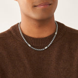 Eine Person trägt einen braunen Pullover und die Fossil Halskette - All Stacked Up Bicolor von Fossil, eine silberne Halskette mit rechteckigen Metallakzenten.