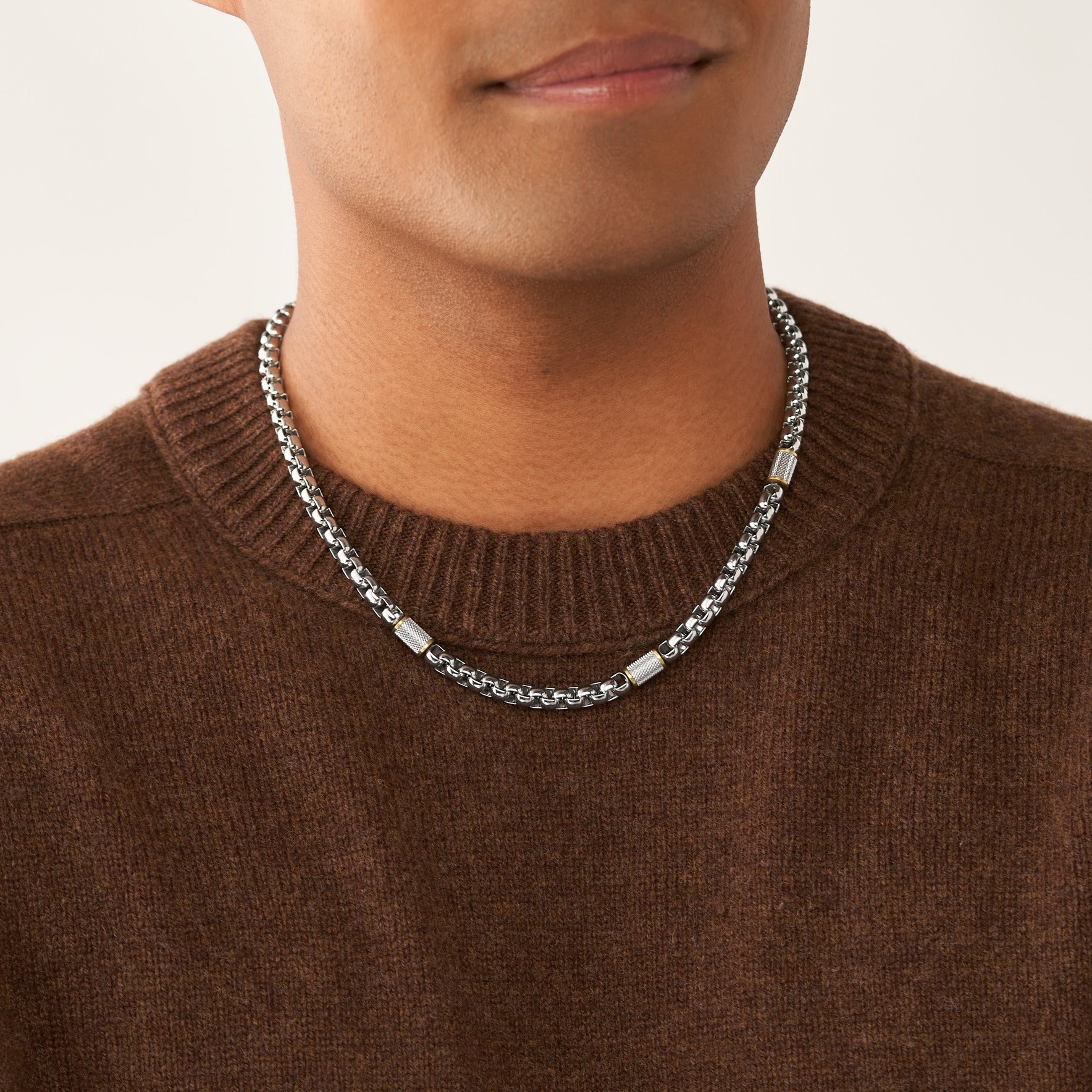 Eine Person trägt einen braunen Pullover und die Fossil Halskette - All Stacked Up Bicolor von Fossil, eine silberne Halskette mit rechteckigen Metallakzenten.