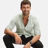 Ein Mann in einem hellgrünen Button-up-Hemd und einer schwarzen Hose lächelt vor einem unifarbenen Hintergrund und trägt die Fynch-Hatton Armbanduhr - The Leather Chrono für einen raffinierten Touch.