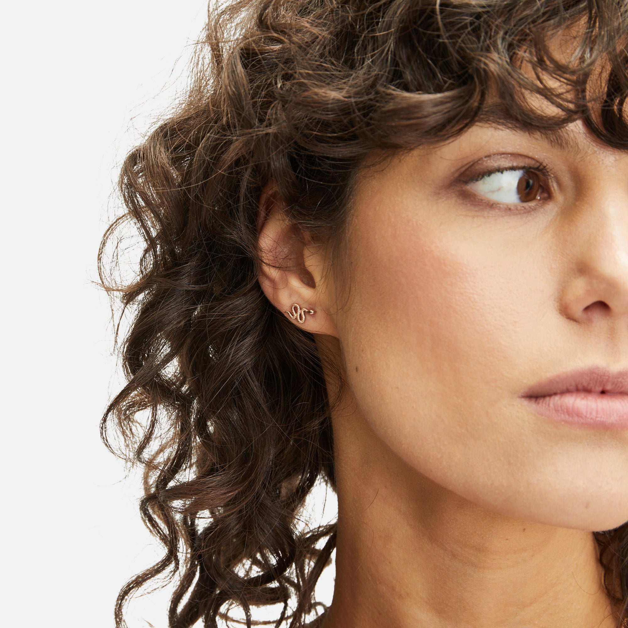 Frau mit lockigem braunem Haar trägt den stilvollen goldenen Ohrstecker "Apple of Eden Ohrstecker - The Stylish Snake", einen zeitlosen Ohrring, der vom Apfel von Eden inspiriert ist, mit Blick nach rechts.