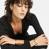 Eine Frau mit lockigem Haar und schwarzem Outfit sitzt mit verschränkten Armen da und präsentiert elegant den Apple of Eden Armreif - The Chic Wave - Slim von Apple of Eden, einen modernen Silberarmreif, der ihrem Look einen schicken Touch verleiht.