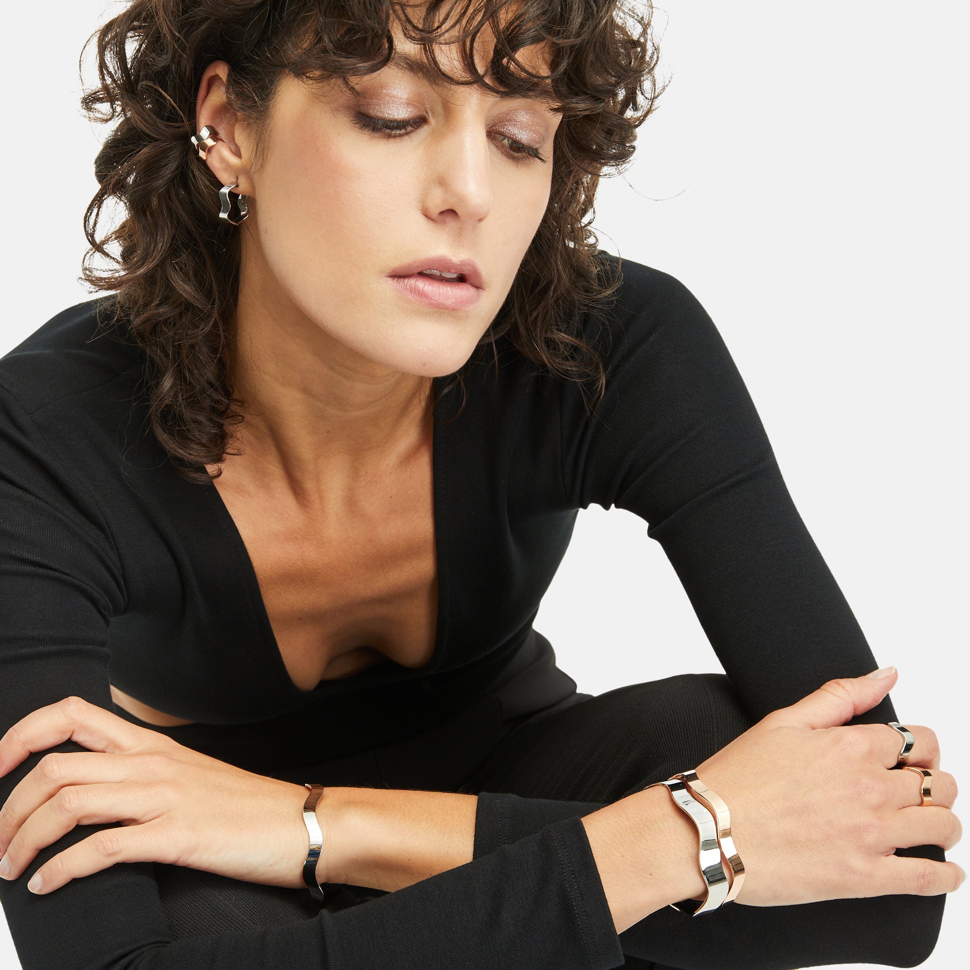 Eine Frau mit lockigem Haar und schwarzem Outfit sitzt mit verschränkten Armen da und präsentiert elegant den Apple of Eden Armreif - The Chic Wave - Slim von Apple of Eden, einen modernen Silberarmreif, der ihrem Look einen schicken Touch verleiht.