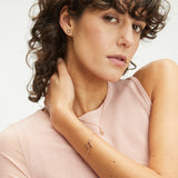 Frau mit lockigem Haar in einem rosa Oberteil trägt einen goldenen Apple of Eden Ohrstecker - The Sparkling Flower sowie eine passende Halskette und ein Armband mit filigranen Details, während sie in die Kamera schaut.