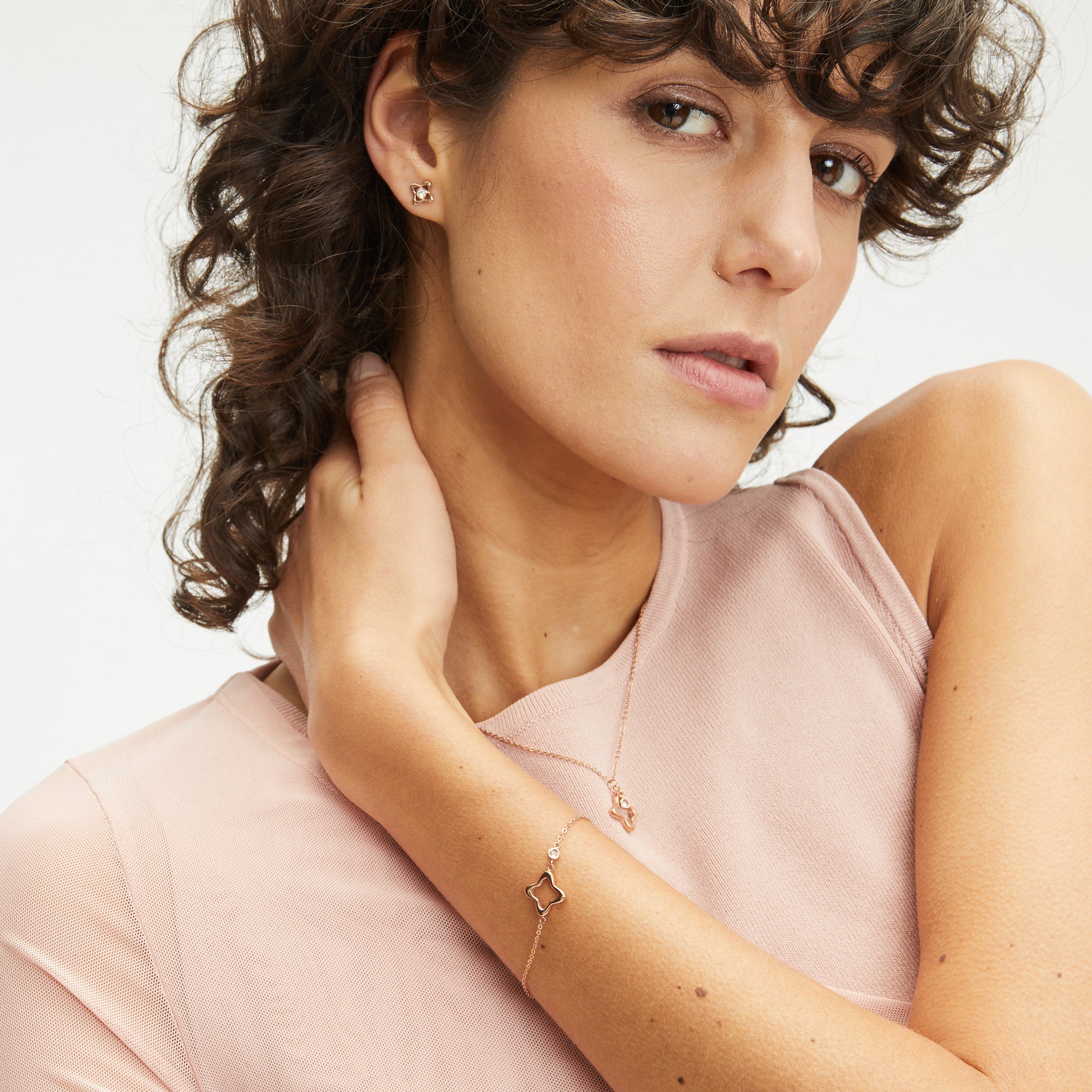 Frau mit lockigem Haar in einem rosa Oberteil trägt einen goldenen Apple of Eden Ohrstecker - The Sparkling Flower sowie eine passende Halskette und ein Armband mit filigranen Details, während sie in die Kamera schaut.