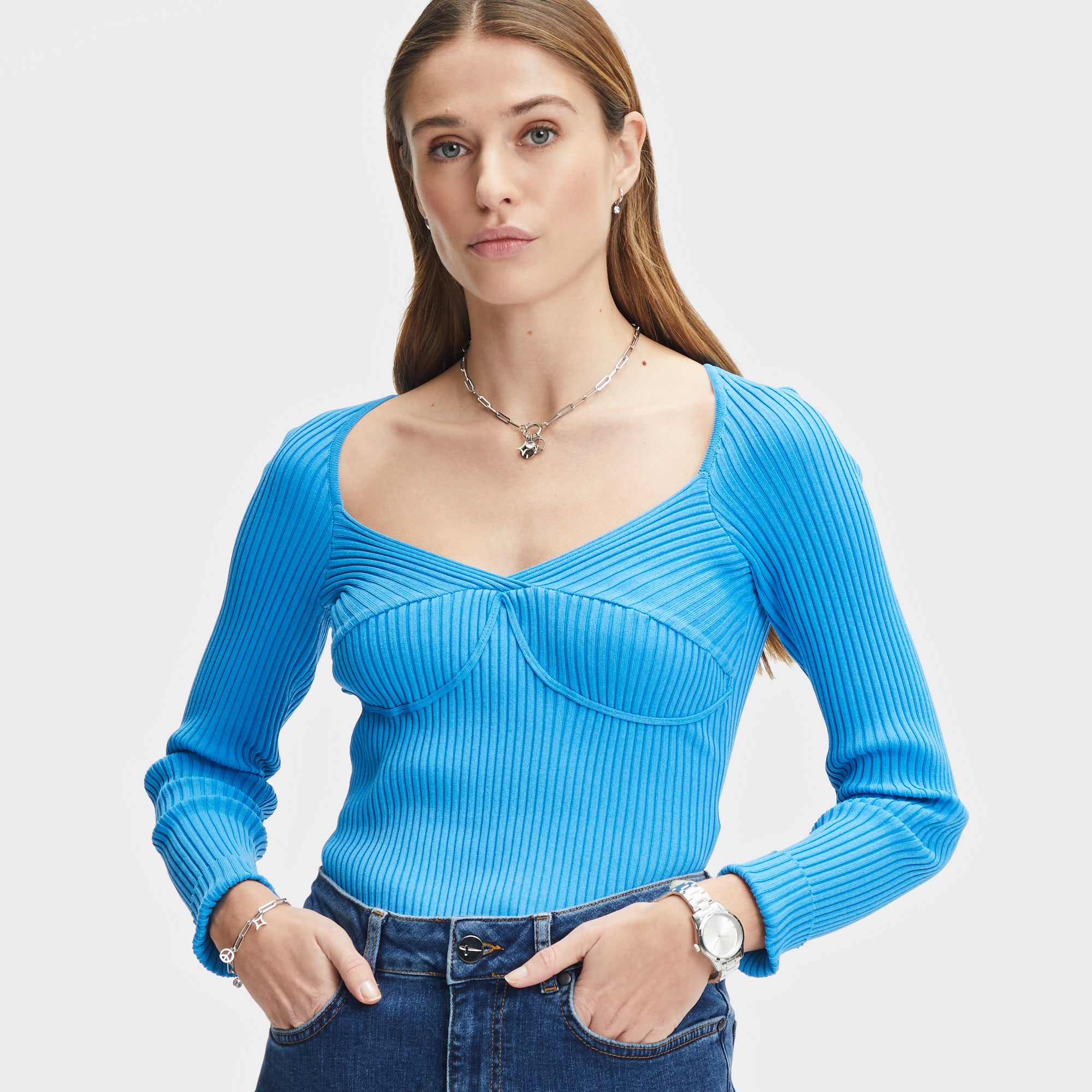 Eine Frau in einem taillierten, langärmeligen blauen Top und Jeans steht vor einem hellen Hintergrund. Ihr Look wird durch das Tamaris Armband - Time to Smile von Tamaris ergänzt.