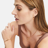 Frau in einem braunen Tank-Top trägt Goldschmuck - Halskette, Ohrringe und Armband - und schaut zur Seite. Ideal zum Verschenken oder um Tamaris Ohrhänger - Glaube, Liebe, Hoffnung von Tamaris elegant zu präsentieren.