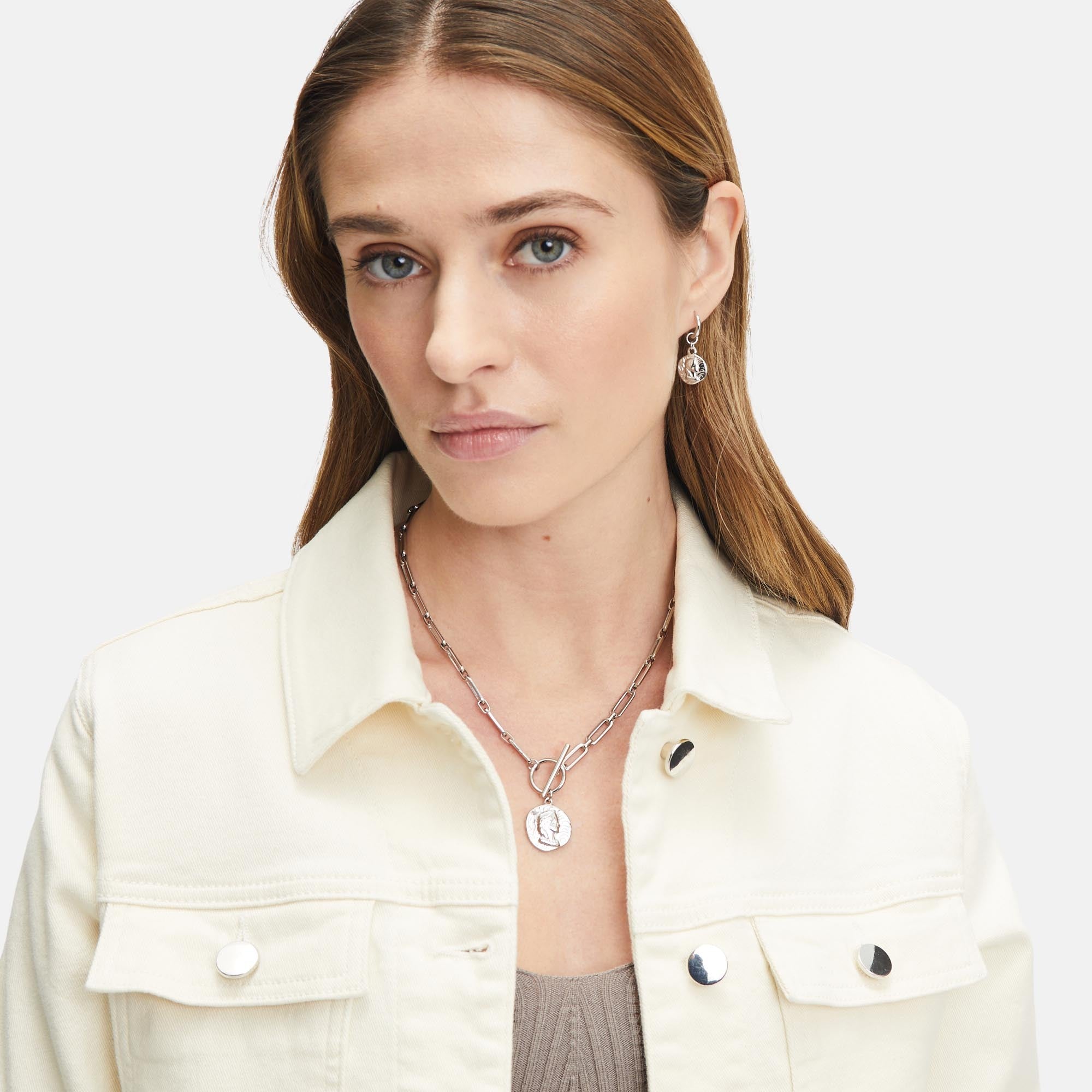 Eine Frau, die eine cremefarbene Jeansjacke und mehrlagigen Silberschmuck, darunter die Creole - Coin Ohrringe von Tamaris, trägt, blickt mit einem neutralen Ausdruck in die Kamera.