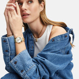 Frau in Jeansjacke und weißem Tanktop mit goldener Uhr und der Tamaris Halskette - Time to Smile von Tamaris, mit Blick zur Seite.
