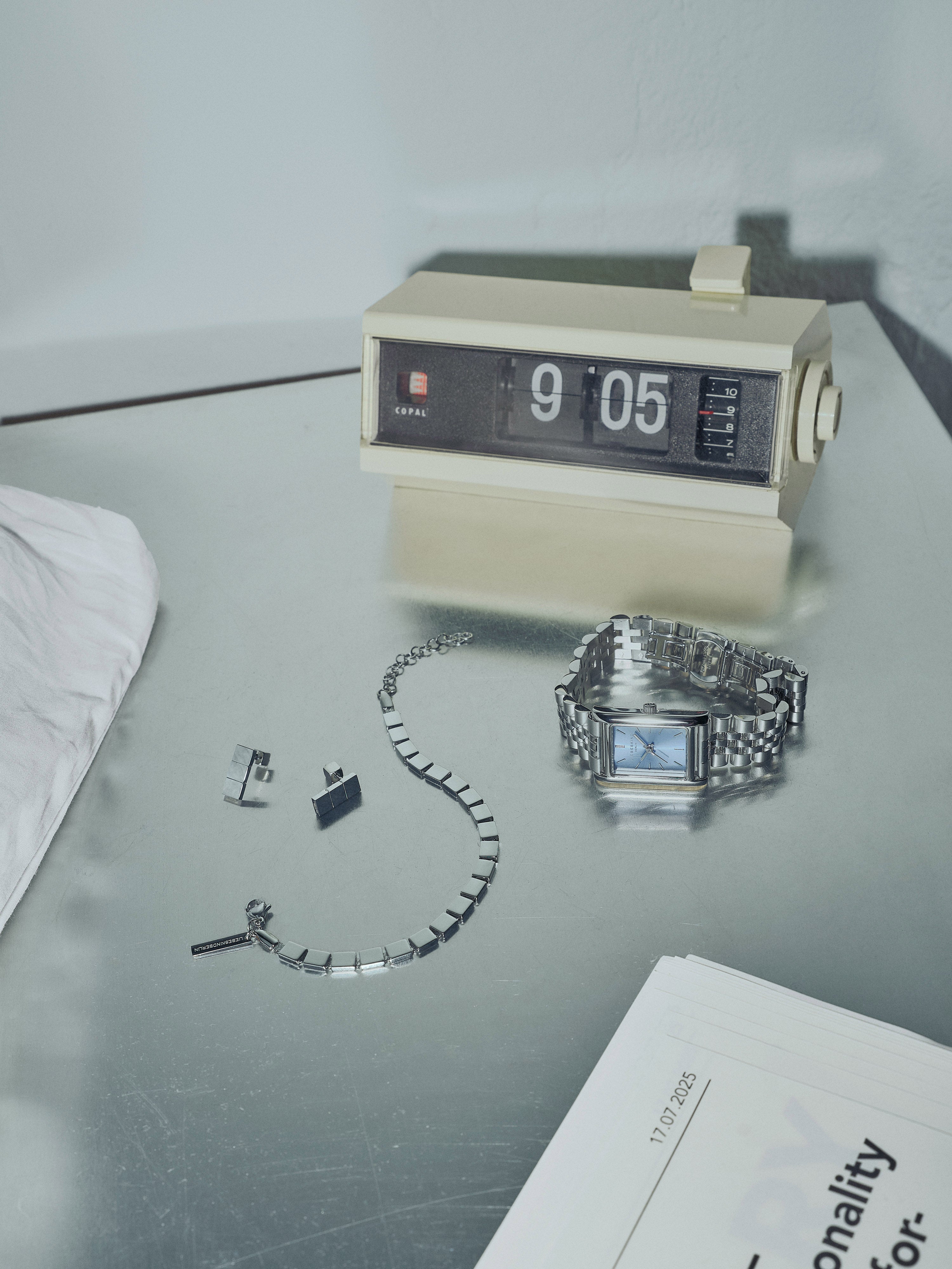 Stilvolle Schmuck- und Uhrenkollektion von Liebeskind Berlin mit markanten, kantigen Designs: eine silberne Armbanduhr mit rechteckigem Zifferblatt, passendes Armband und Ohrringe in geometrischer Form, arrangiert neben einer Retro-Klappzahlen-Uhr auf einem silbernen Tisch.