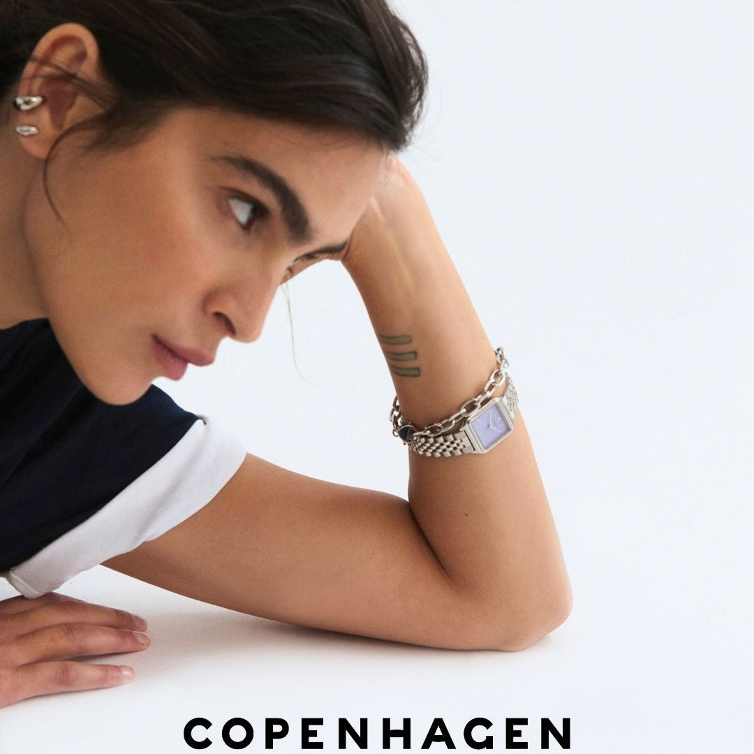 Frau mit dunklen Haaren stützt sich auf den Arm, zeigt eine Uhr und Tätowierungen, darunter der Text Copenhagen Studios.