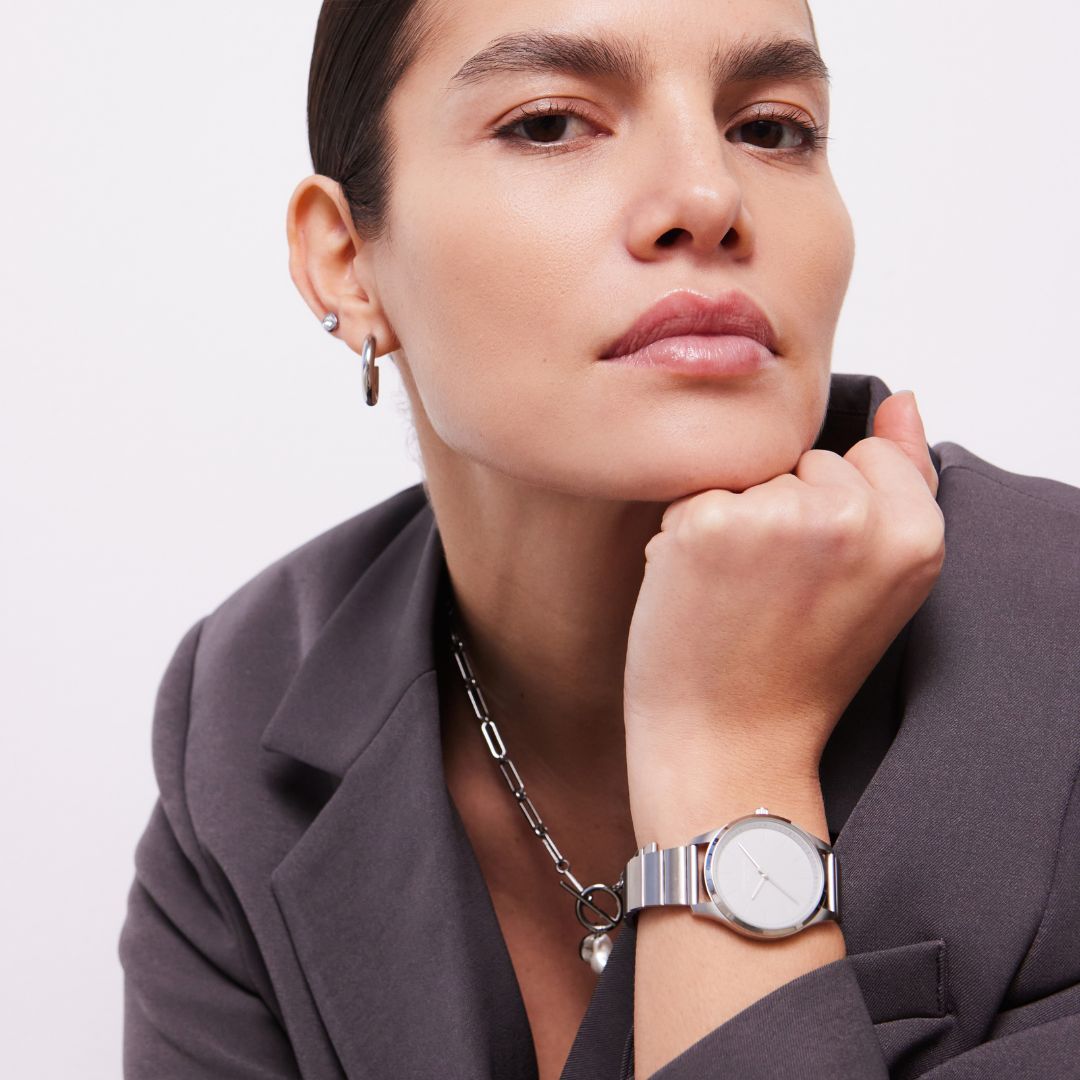 Frau im grauen Blazer stützt das Kinn auf die Hand und trägt silberne Ohrringe, Kette und Uhr.