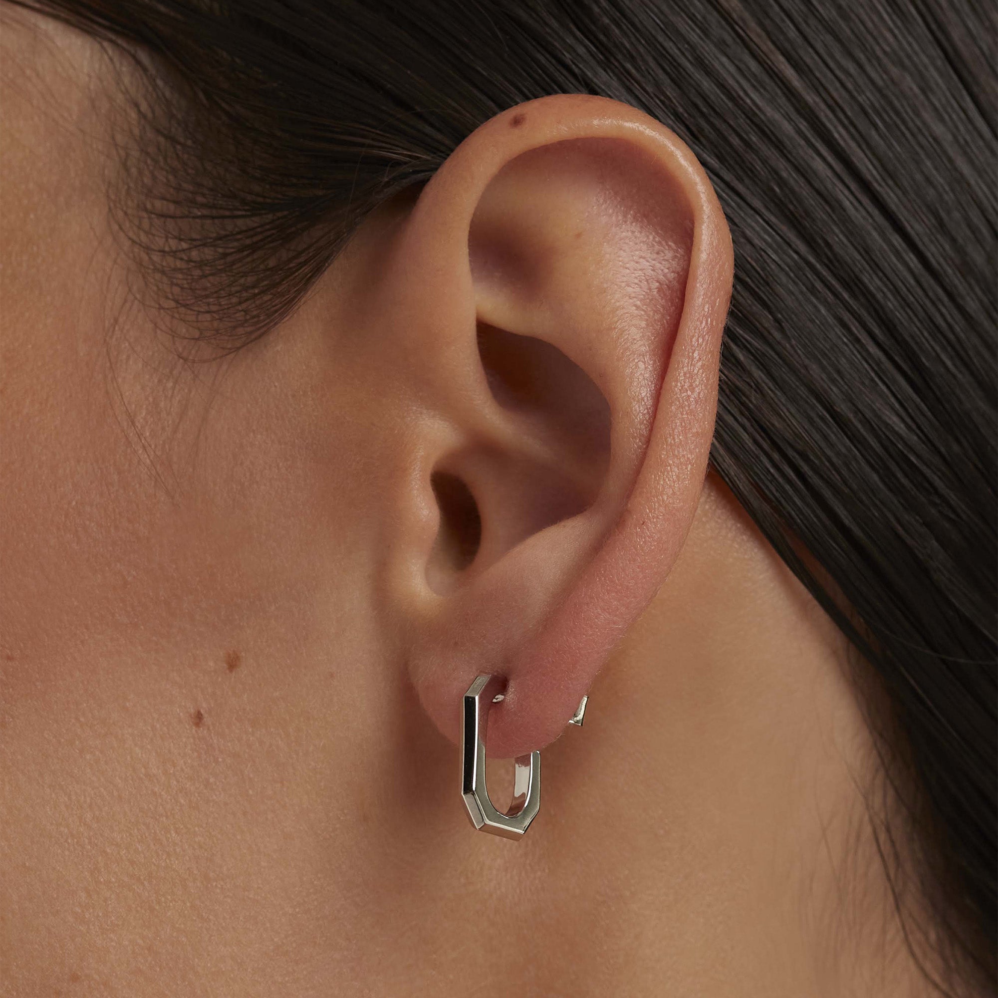 Nahaufnahme des Ohrs einer Person, die PDPaola Ohrhänger - The Chain, einen geometrischen silbernen Ohrring von PDPaola trägt, mit glattem braunem Haar, das nach hinten gezogen ist.