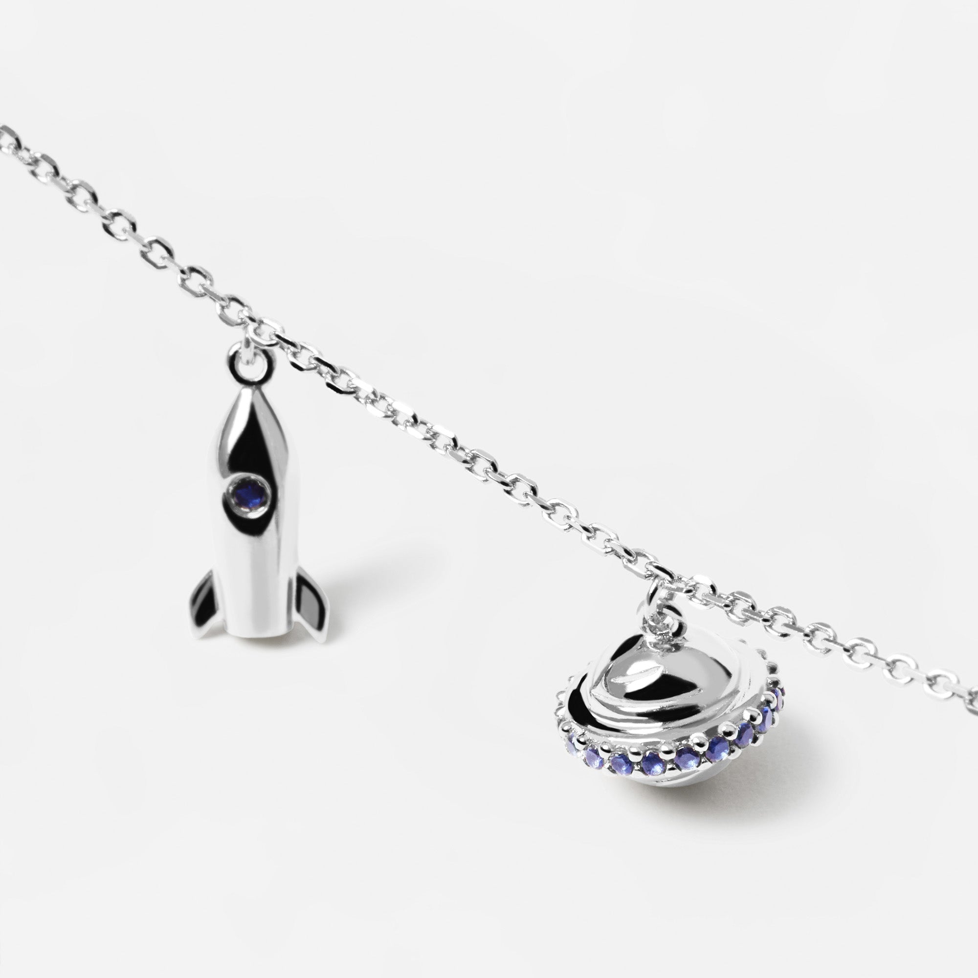 Die PDPaola Halskette - The Mini Charms zeigt Raketen- und Ufo-Anhänger aus Silber, beide mit kleinen blauen Edelsteinen verziert, präsentiert auf weißem Hintergrund.