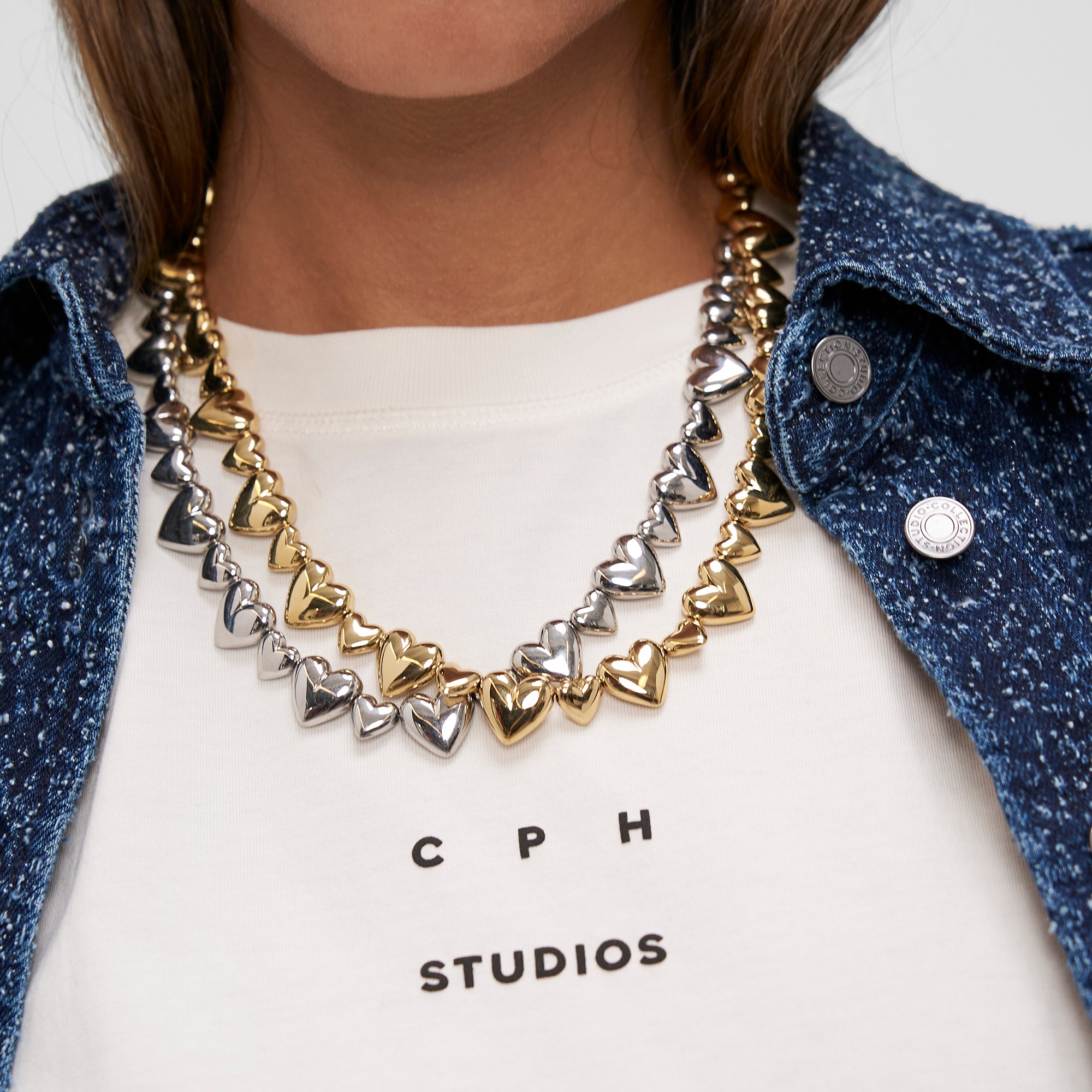 Nahaufnahme von zwei metallischen Herzketten, eine goldene und eine silberne, gestylt auf einem weißen Hemd - und die COPENHAGEN STUDIOS Halskette - Puffy Hearts by Copenhagen Studios mit ihrem eleganten Herzkarabiner-Detail hervorhebend.